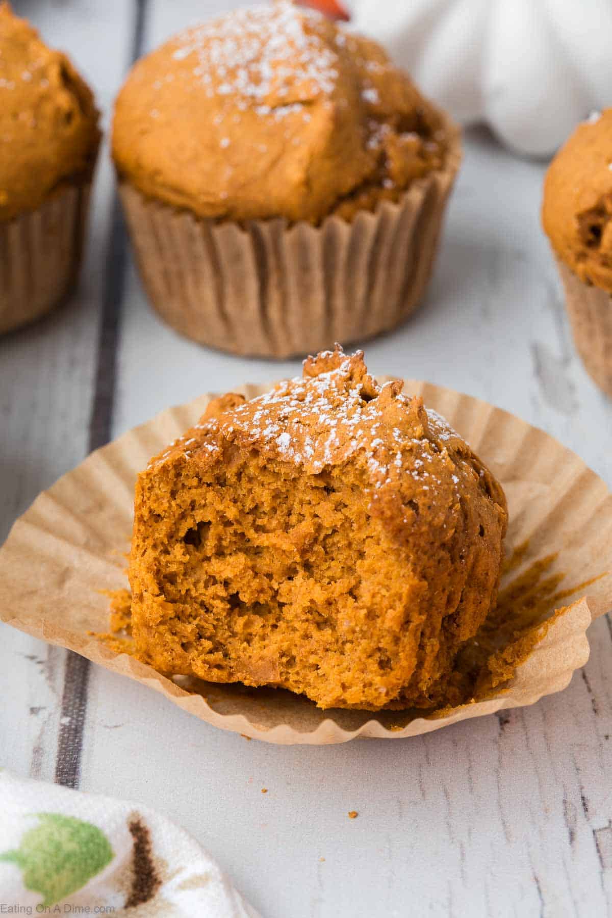 A close-up of a 2 Ingredient Pumpkin Muffin with a bite taken out sits partially unwrapped from its paper liner. The muffin is sprinkled with powdered sugar, adding a touch of sweetness. Two more intact muffins are seen in the background on a white wooden surface, inviting you to indulge.