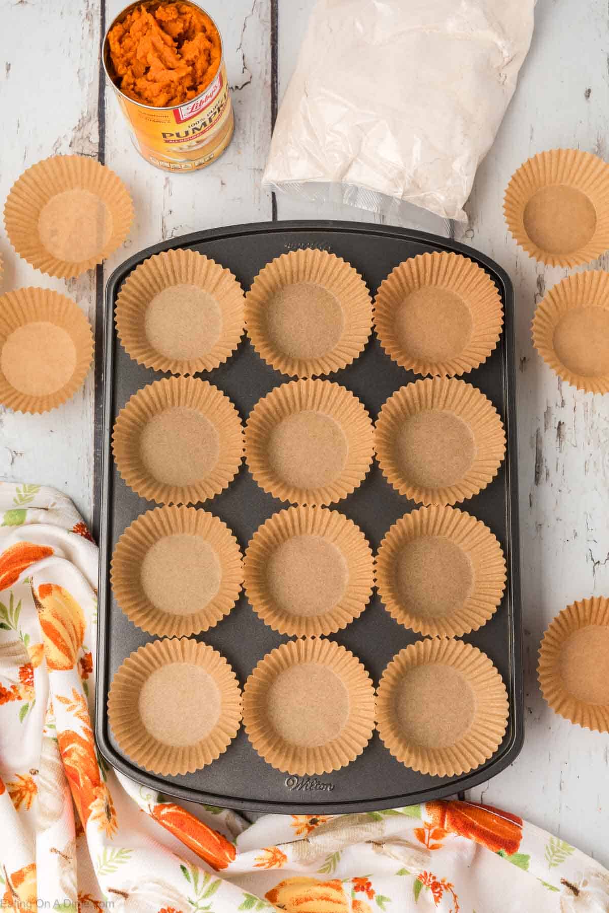 A muffin tin filled with brown paper cupcake liners is centered on a light wooden table. Surrounding the tin are a can of pumpkin puree, a bag of flour, scattered cupcake liners, and a floral-patterned cloth. The baking setup suggests preparation for 2 Ingredient Pumpkin Muffins.