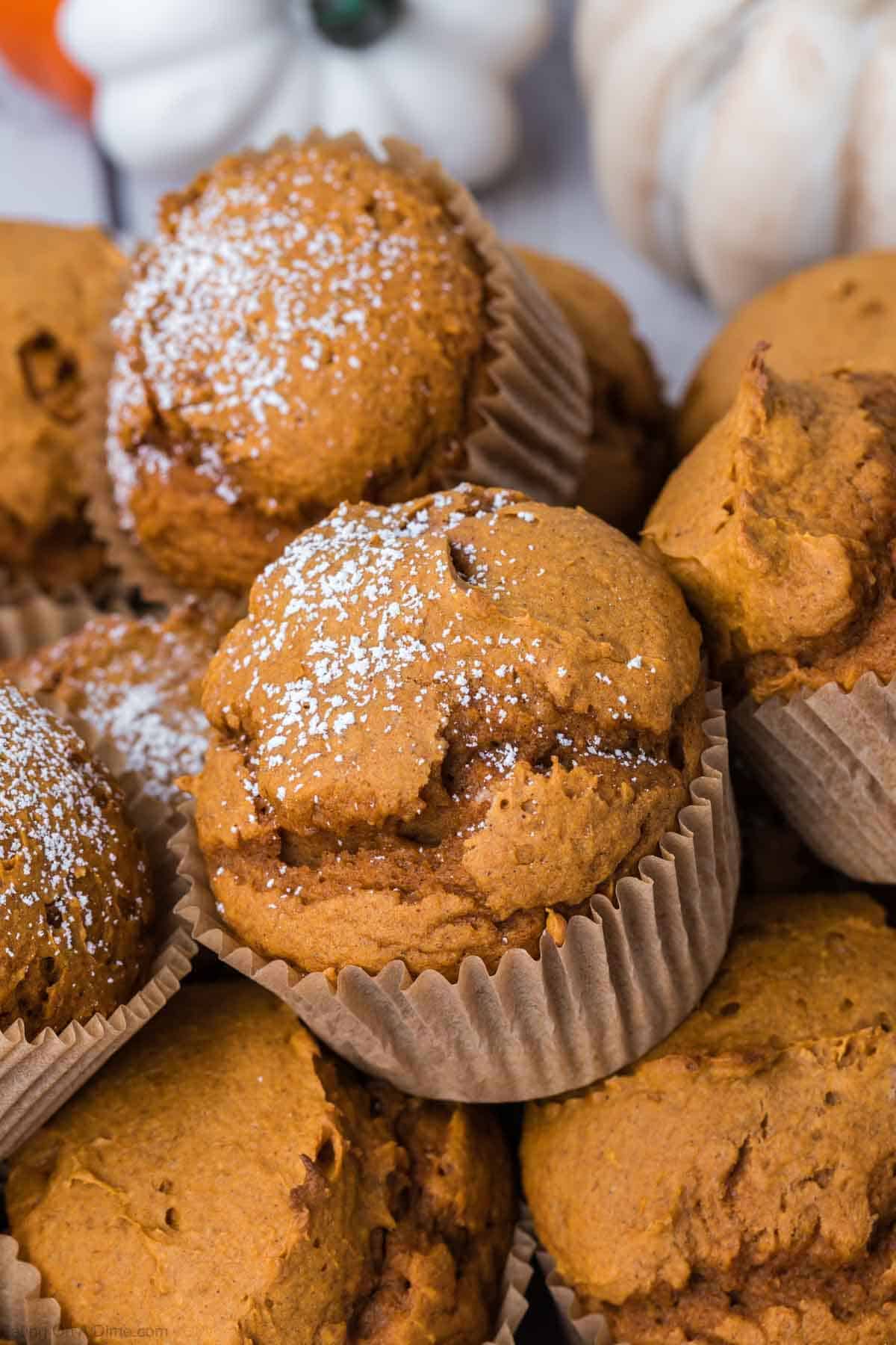 A stack of freshly baked 2 Ingredient Pumpkin Muffins with light dustings of powdered sugar on top. The muffins are housed in rustic paper liners, giving them a homemade and cozy appearance. Additionally, there are white pumpkins in the blurred background, enhancing the autumnal feel.