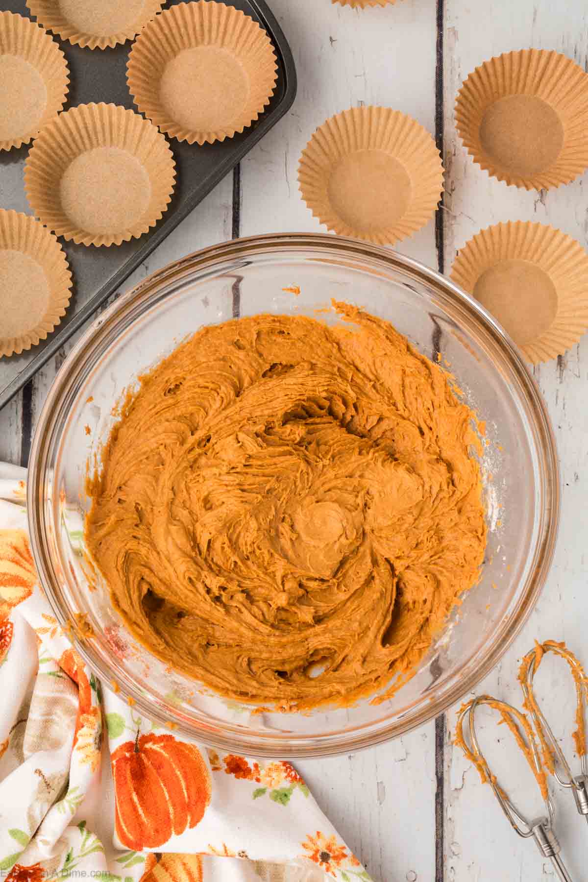 A glass mixing bowl filled with 2 Ingredient Pumpkin Muffins batter sits on a white wooden table. Surrounding the bowl are scattered empty brown muffin liners and a tray with liners set in it. A floral dish towel and a whisk complete the cozy baking scene.