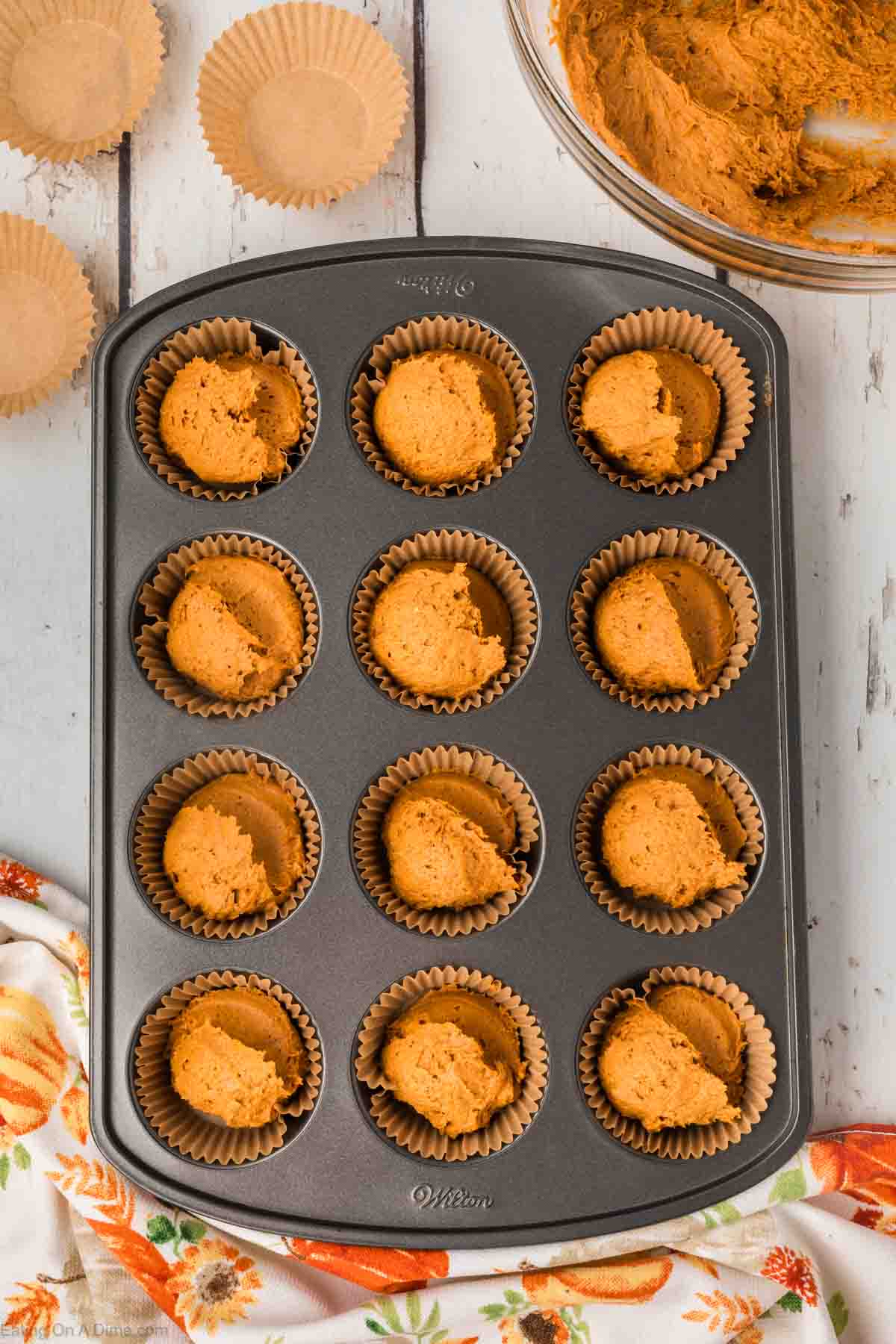 A muffin tray lined with beige paper cups, each filled with dollops of orange batter for 2 Ingredient Pumpkin Muffins, is placed on a white wooden surface. A bowl with remaining orange batter and additional paper cups are nearby. The corner of a patterned cloth with orange and green designs is visible.