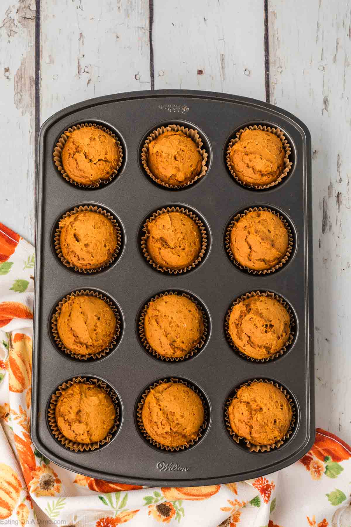A muffin tin filled with twelve freshly baked 2 ingredient pumpkin muffins, each in a brown paper liner. The tin is placed on a rustic wooden surface, and a colorful orange and white kitchen towel is partially visible at the bottom left corner of the image.
