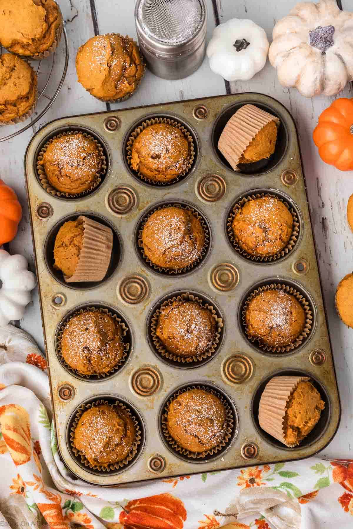A muffin tray filled with 2 ingredient pumpkin muffins sits on a white wooden table. Some muffins are in paper liners, and others are without. The muffins are topped with powdered sugar. Small decorative pumpkins surround the tray, and a floral cloth is partly visible.