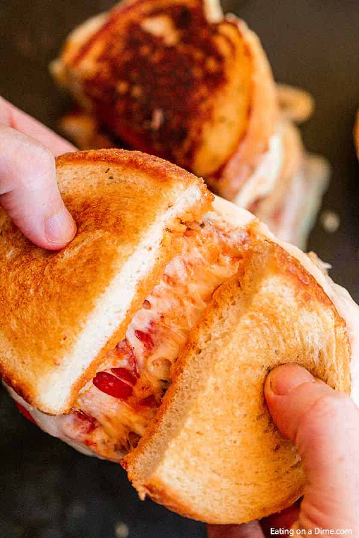 A person holds a Pizza Grilled Cheese Sandwich with hands, pulling the two halves apart to reveal the melted cheese and pieces of tomato inside. Another grilled sandwich is blurred in the background. The image's bottom right corner has the text "Eating on a Dime.com.