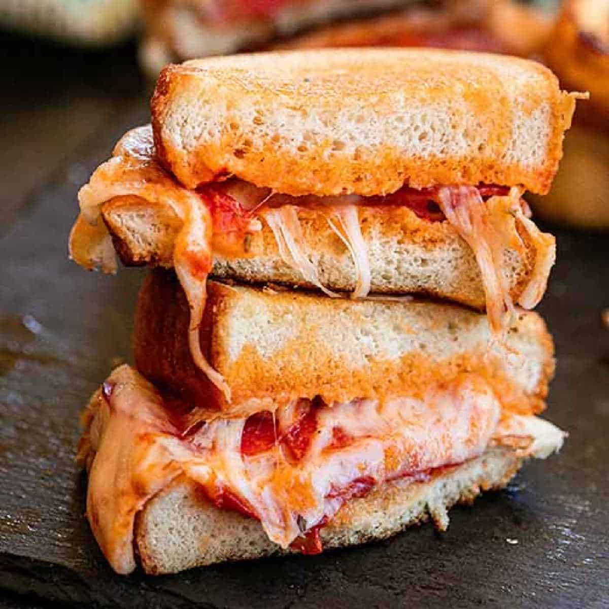 Close-up of a pizza grilled cheese sandwich overflowing with melted cheese and bits of tomato. The sandwich is cut in half, with the two halves stacked on top of each other, showing the gooey, cheesy filling oozing out between the toasted bread slices.