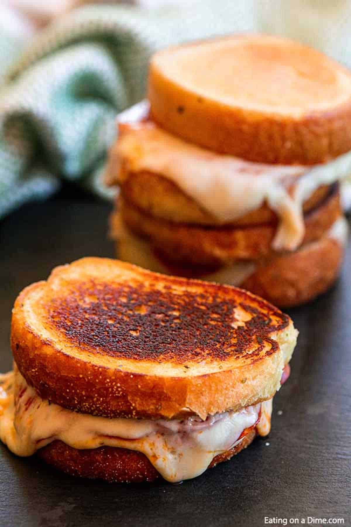 A close-up shot of two pizza grilled cheese sandwiches with golden brown, toasted bread and melted cheese oozing out. A stack of more grilled cheese sandwiches is visible in the background, with a green and white cloth partially visible on a dark surface.