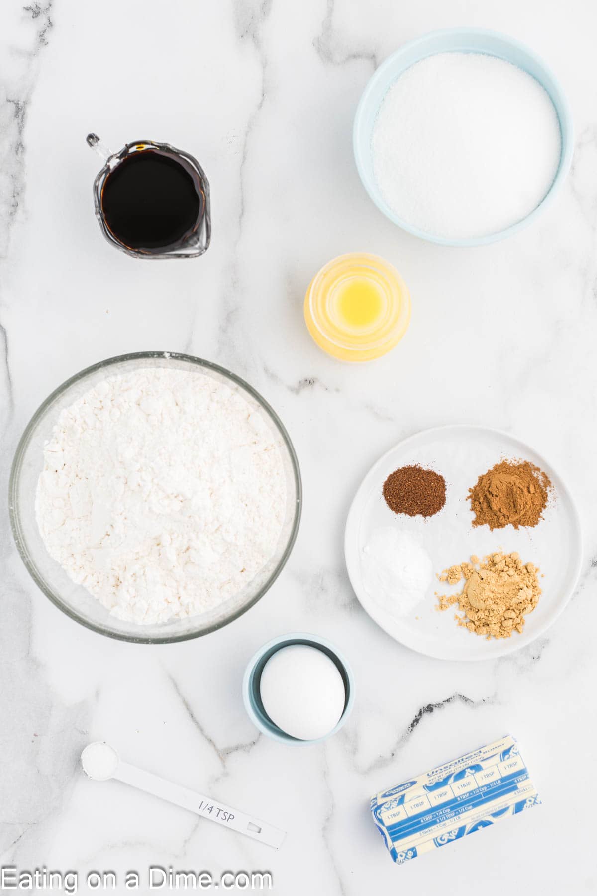 Baking ingredients for chewy gingerbread cookies are laid out on a marble surface: a bowl of flour, a bowl of sugar, a small bowl with yellow liquid, a small black liquid container, a plate with spices, and buttery essentials waiting to transform into deliciousness.