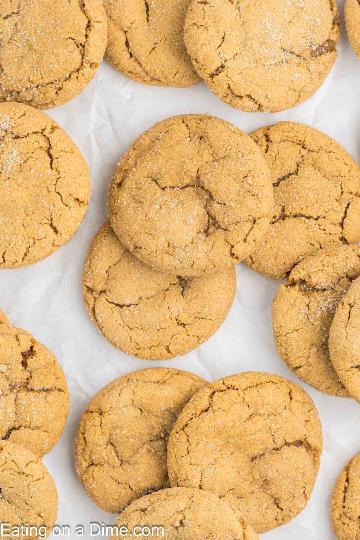 A close-up of several golden-brown chewy gingerbread cookies with crackled surfaces, dusted with a light layer of sugar. They are arranged on a piece of parchment paper.