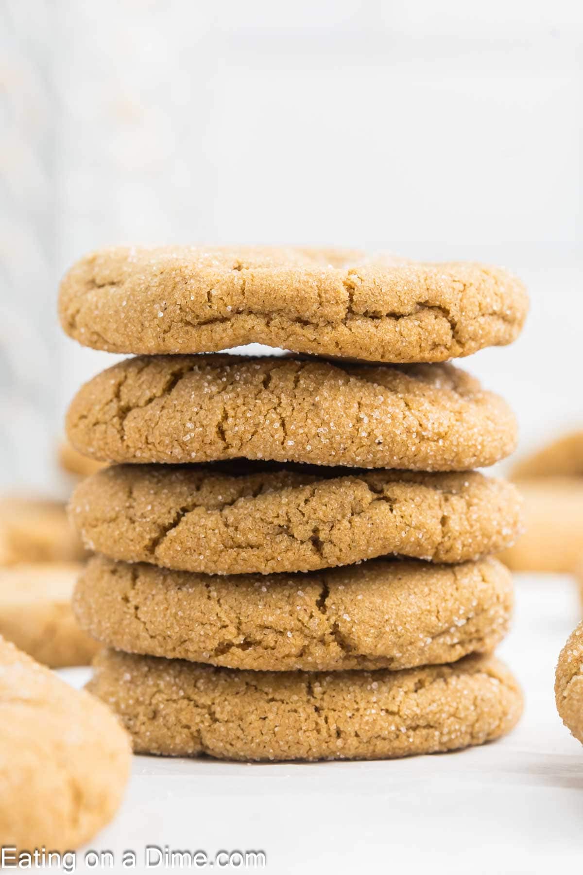 A close-up of a stack of five sugar-coated, golden-brown cookies with a cracked texture sits on a white surface. These chewy gingerbread cookies have other delectable treats blurred in the background.