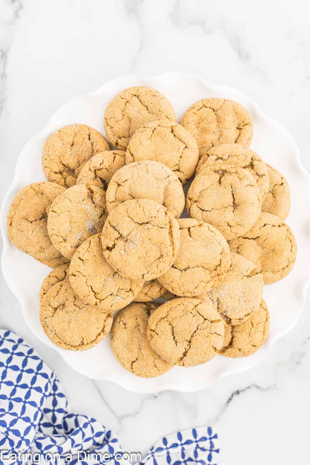 A plate full of freshly baked, chewy gingerbread cookies is stacked neatly on a white plate. The cookies have a crinkled surface and a light dusting of sugar. A blue and white patterned cloth peeks from the corner against a marble background.