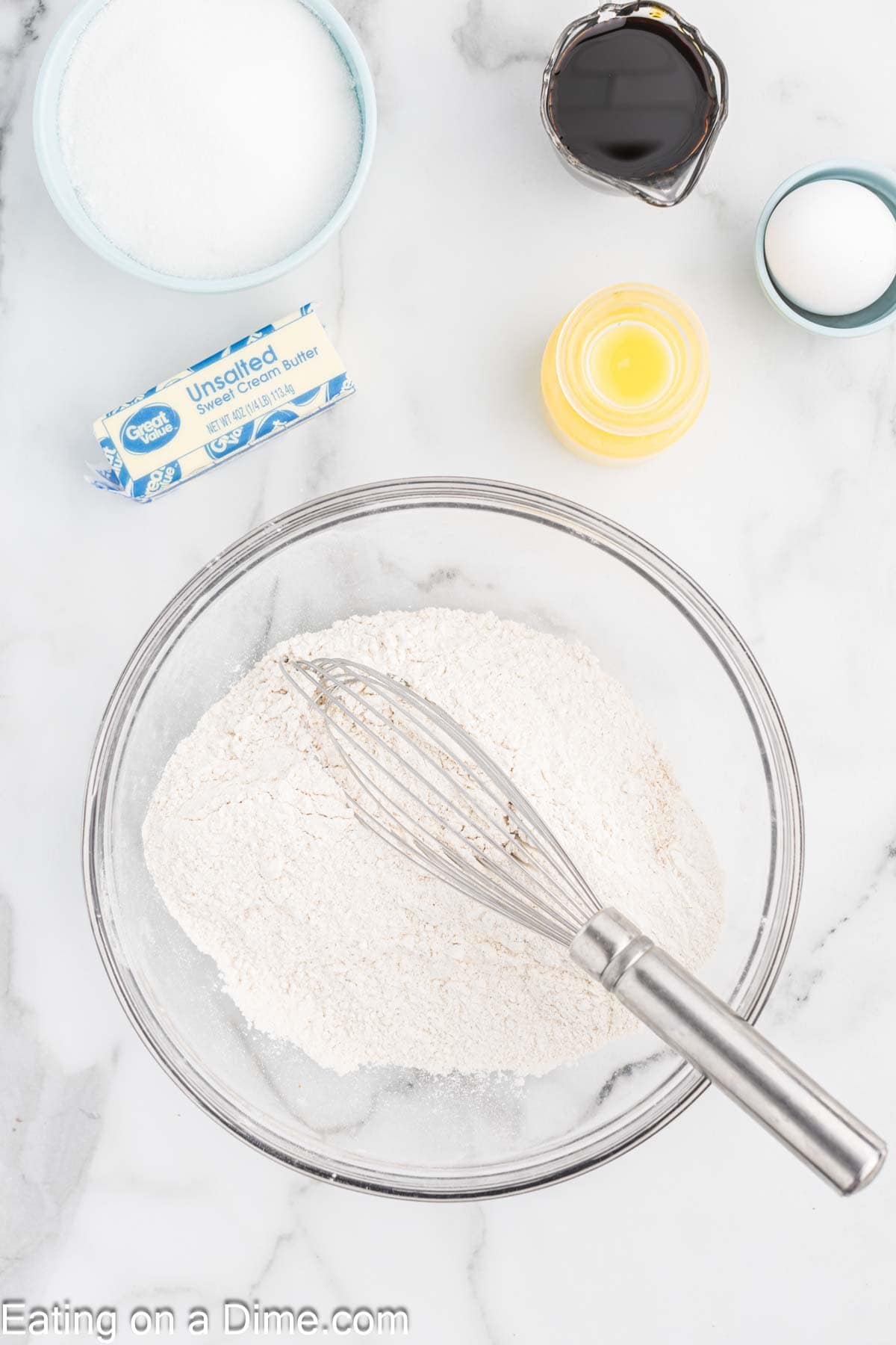 A baking preparation scene for chewy gingerbread cookies features a whisk in a glass bowl of flour. Surrounding ingredients include a stick of unsalted butter, sugar, a small bowl of milk, and a cup of molasses on the white marble surface.