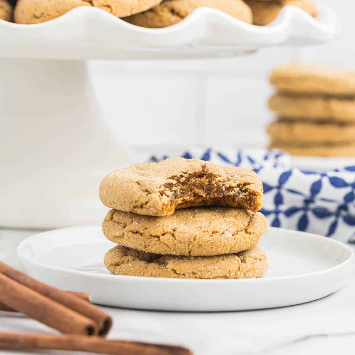 A close-up of three stacked chewy gingerbread cookies on a white plate, with the top cookie broken to reveal a delightful, soft interior. The background features more cookies on a raised stand and a blue patterned cloth. Two cinnamon sticks are in the foreground.