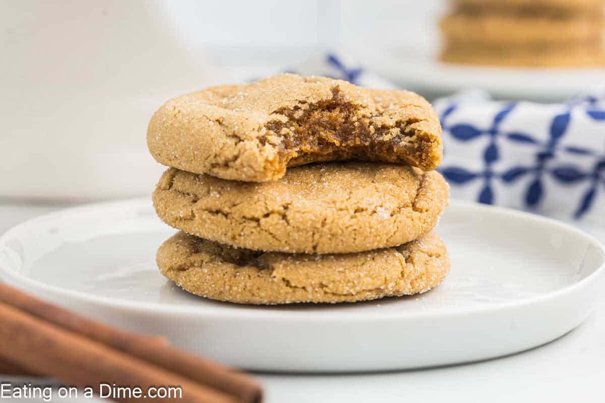 Three stacked chewy gingerbread cookies on a plate, with the top cookie showing a bite taken out of it. A blue and white napkin is in the background, and cinnamon sticks are visible on the side.