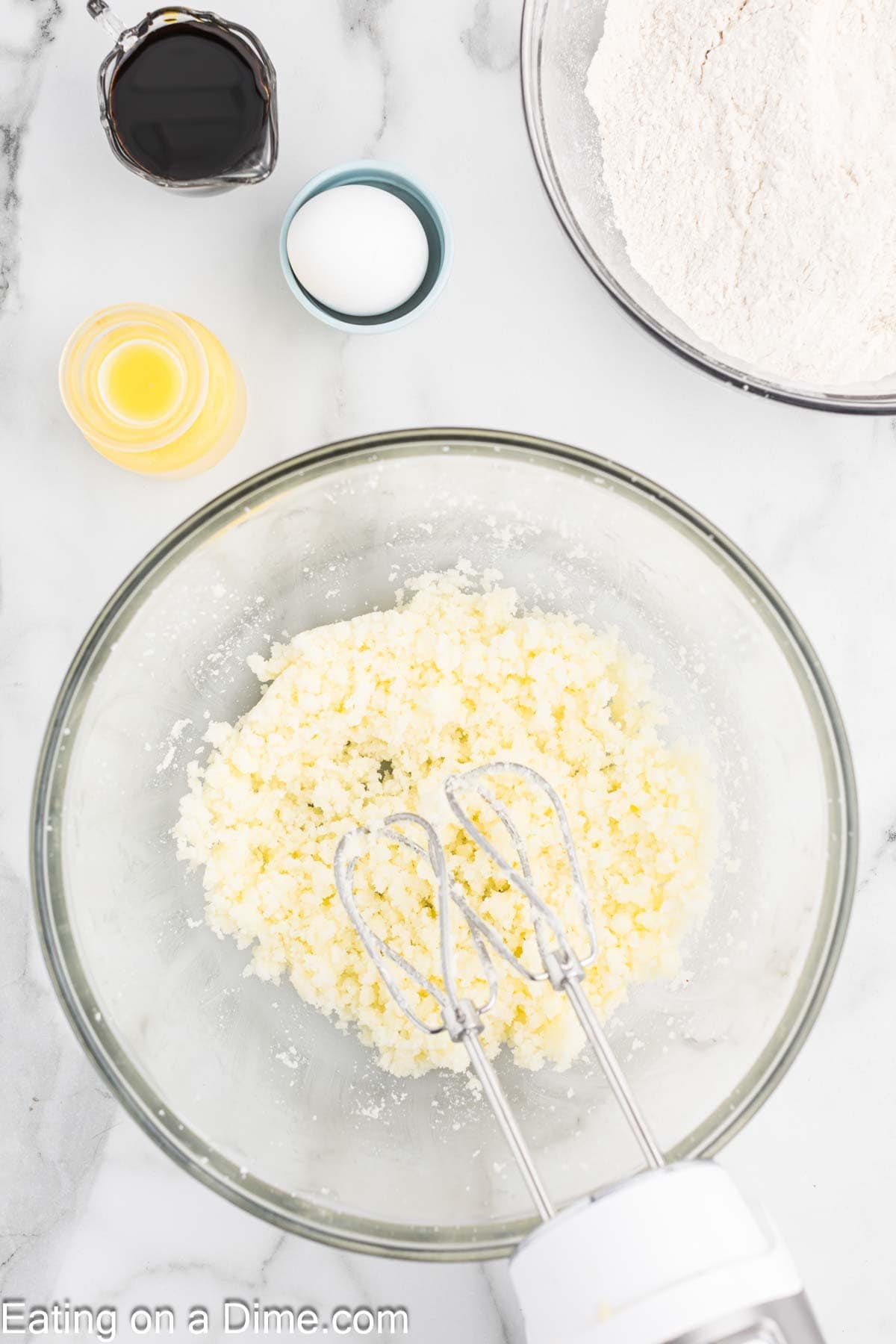 A glass bowl with creamed butter and sugar sits on a marble countertop, surrounded by small bowls of vanilla extract, salt, flour, and orange juice—perfect for making chewy gingerbread cookies. A hand mixer is poised in the creamy mixture, ready for action.