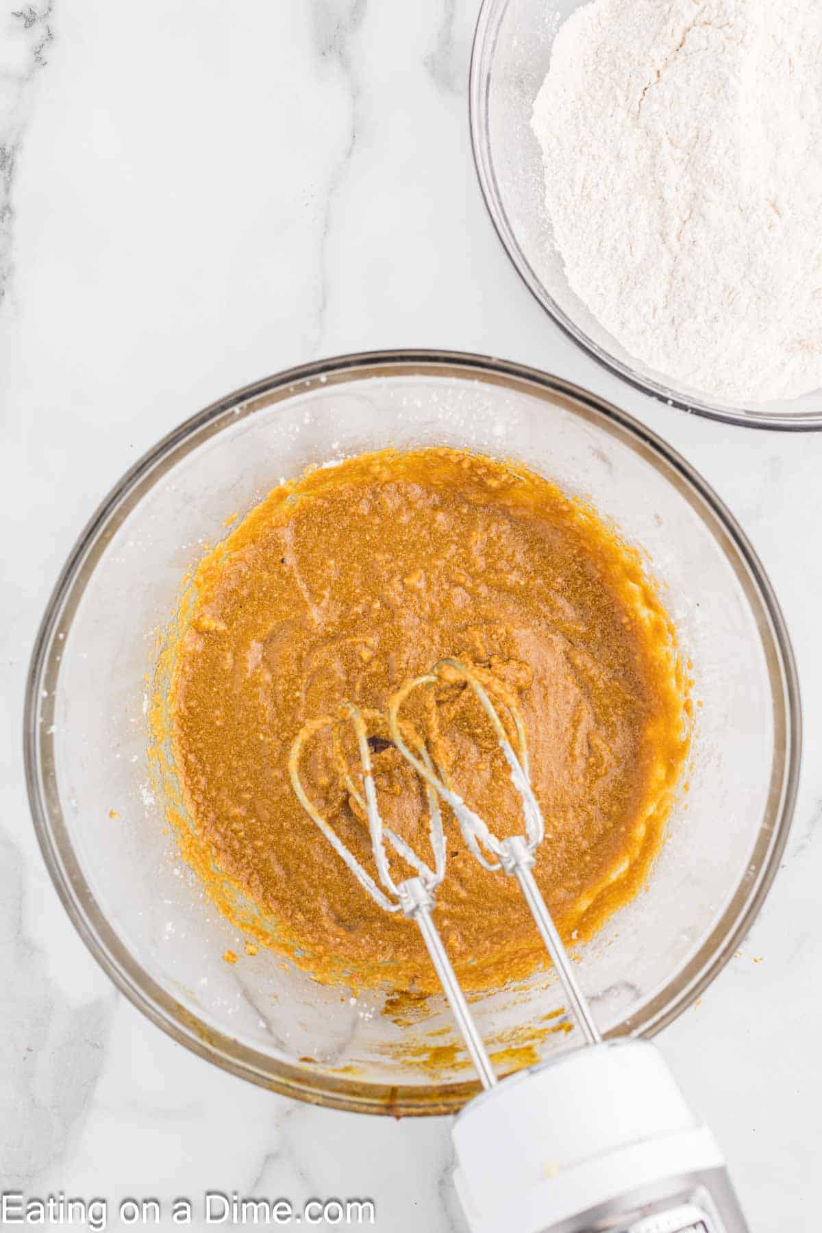 A glass mixing bowl filled with creamy, light brown batter for chewy gingerbread cookies sits alongside an electric mixer. In the background, a separate bowl holds a white, flour-like substance. The scene unfolds on a sleek white marble countertop.