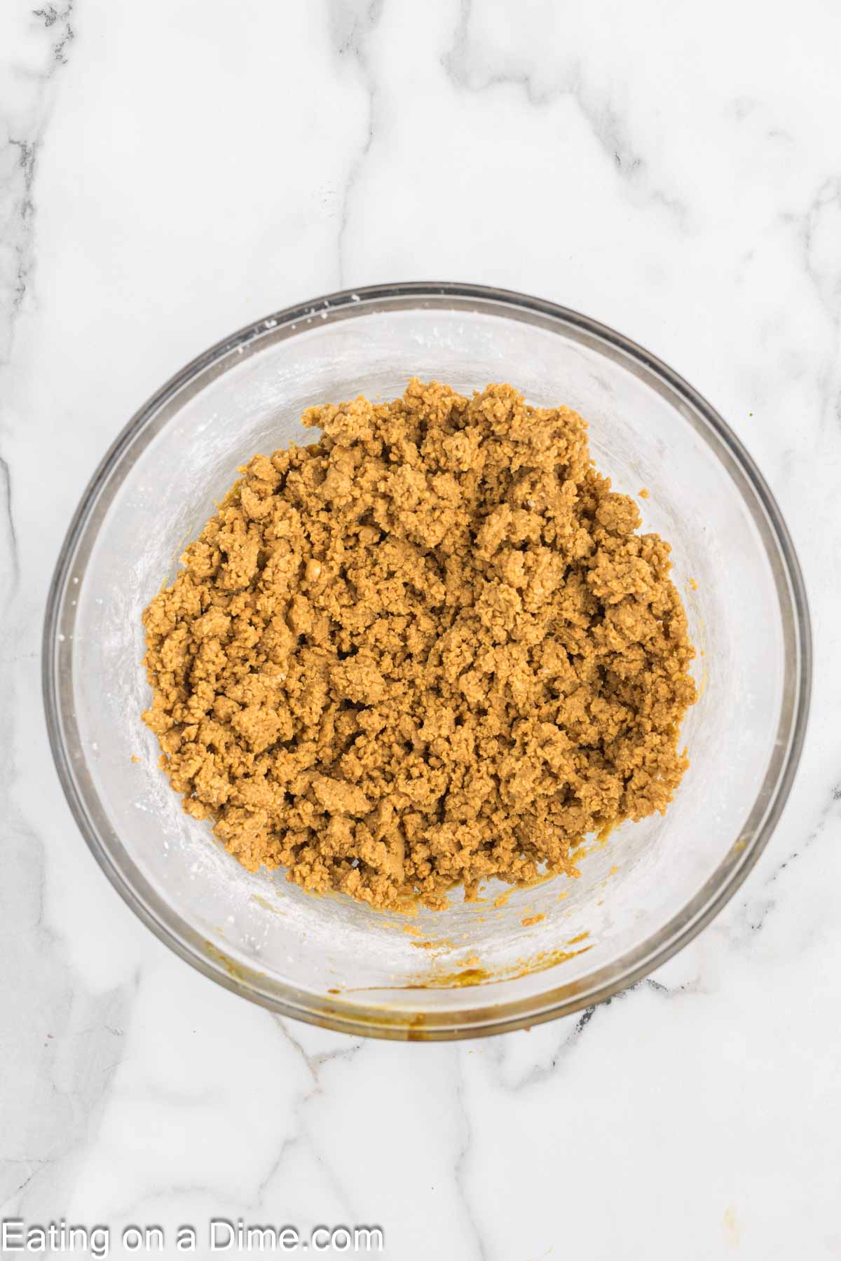 A glass bowl filled with a crumbly brown mixture on a white marble surface hints at the beginning of chewy gingerbread cookies. The mixture appears to be cookie dough, ready to be transformed into warm, spiced delights.