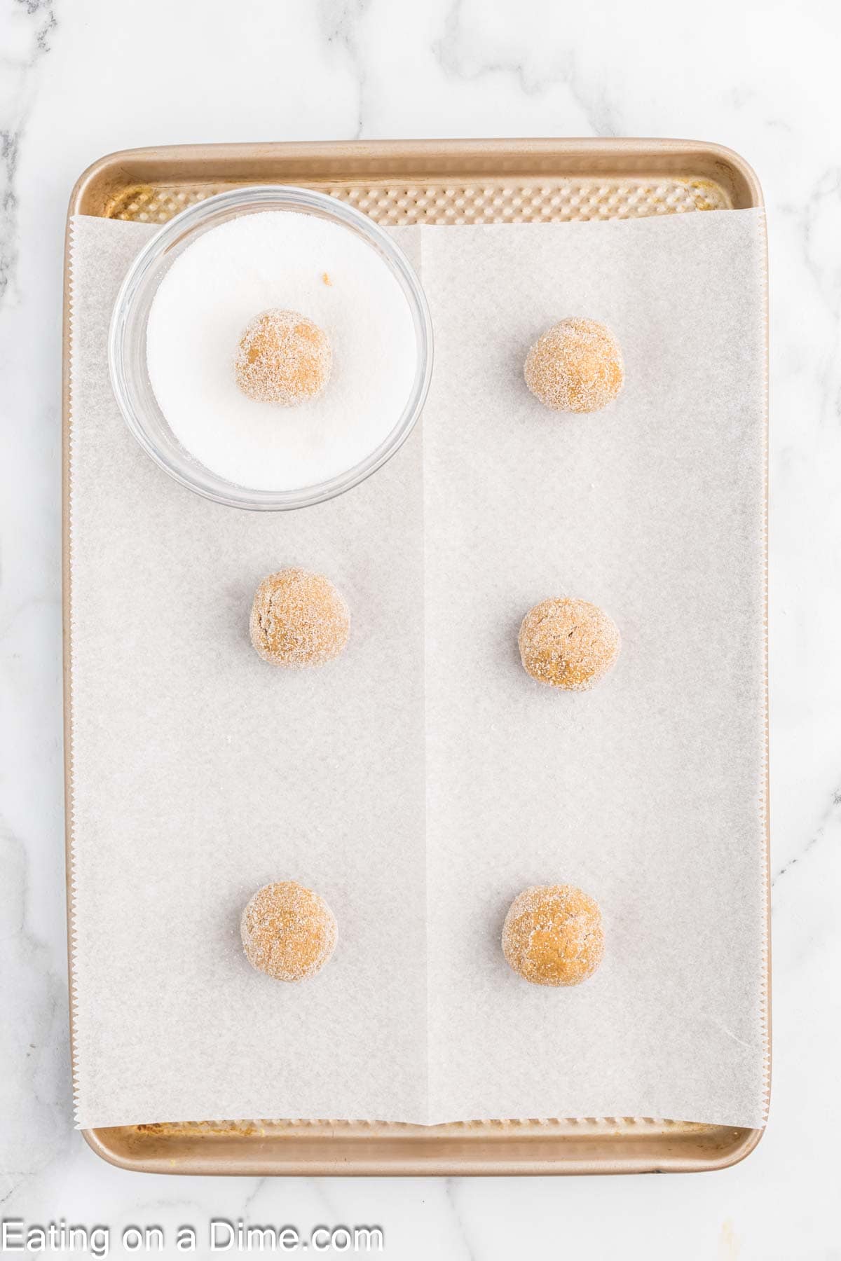 A baking sheet lined with parchment paper holds six sugar-coated cookie dough balls, promising chewy gingerbread cookies to come. A small bowl of sugar sits invitingly on the sheet, ready for the sprinkle before baking. The surface is a light marble pattern, adding elegance to the preparation scene.