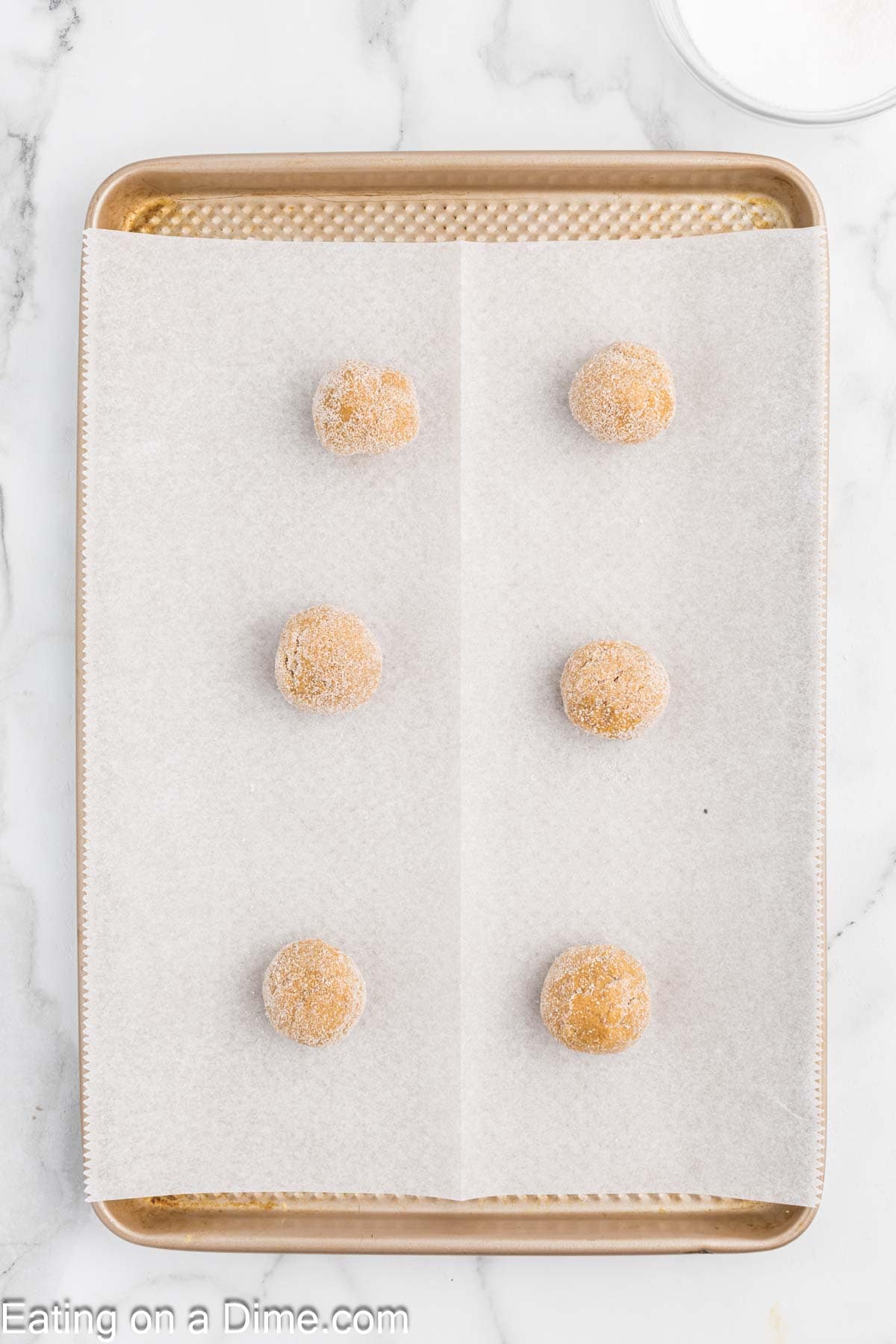 A baking sheet lined with parchment paper holds six evenly spaced dough balls of chewy gingerbread cookies coated in sugar, ready to be baked. The backdrop is a white marble countertop.