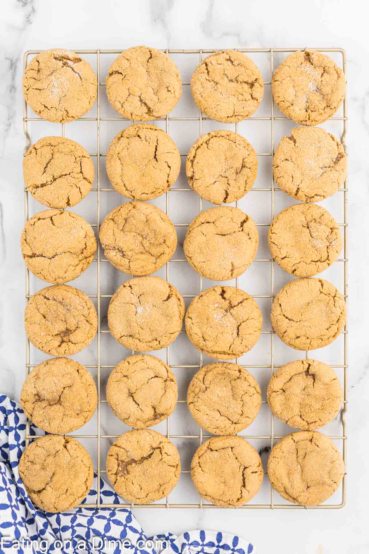 A grid of 24 freshly baked chewy gingerbread cookies with a golden-brown, cracked surface, cooling on a wire rack placed on a marble countertop. A blue and white patterned cloth is partially visible in the bottom left corner.