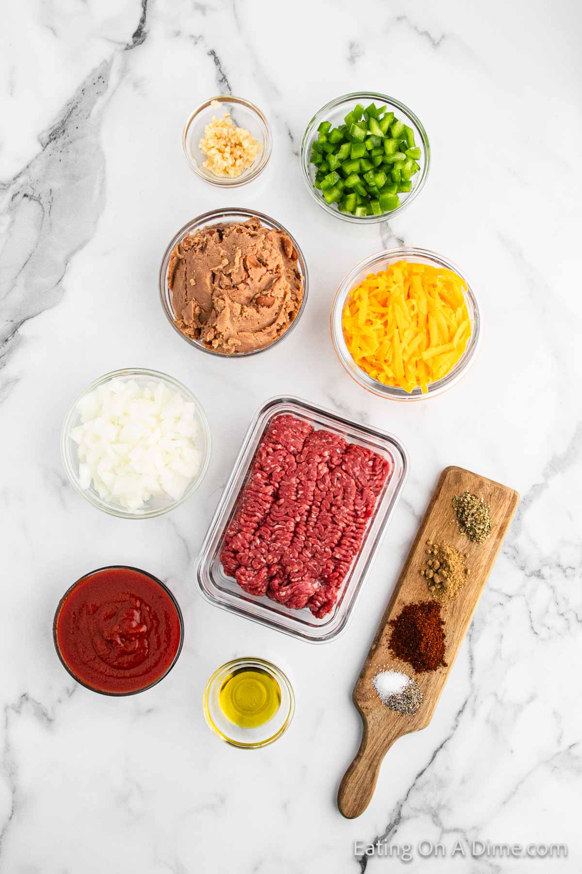 A top-view of taco ingredients on a marble surface showcases everything needed for Beef Chimichangas. Ground beef, shredded cheese, chopped onions, green bell peppers, refried beans, minced garlic, and tomato sauce are arranged in bowls. A wooden board holds various spices. A small bowl of olive oil is also present.