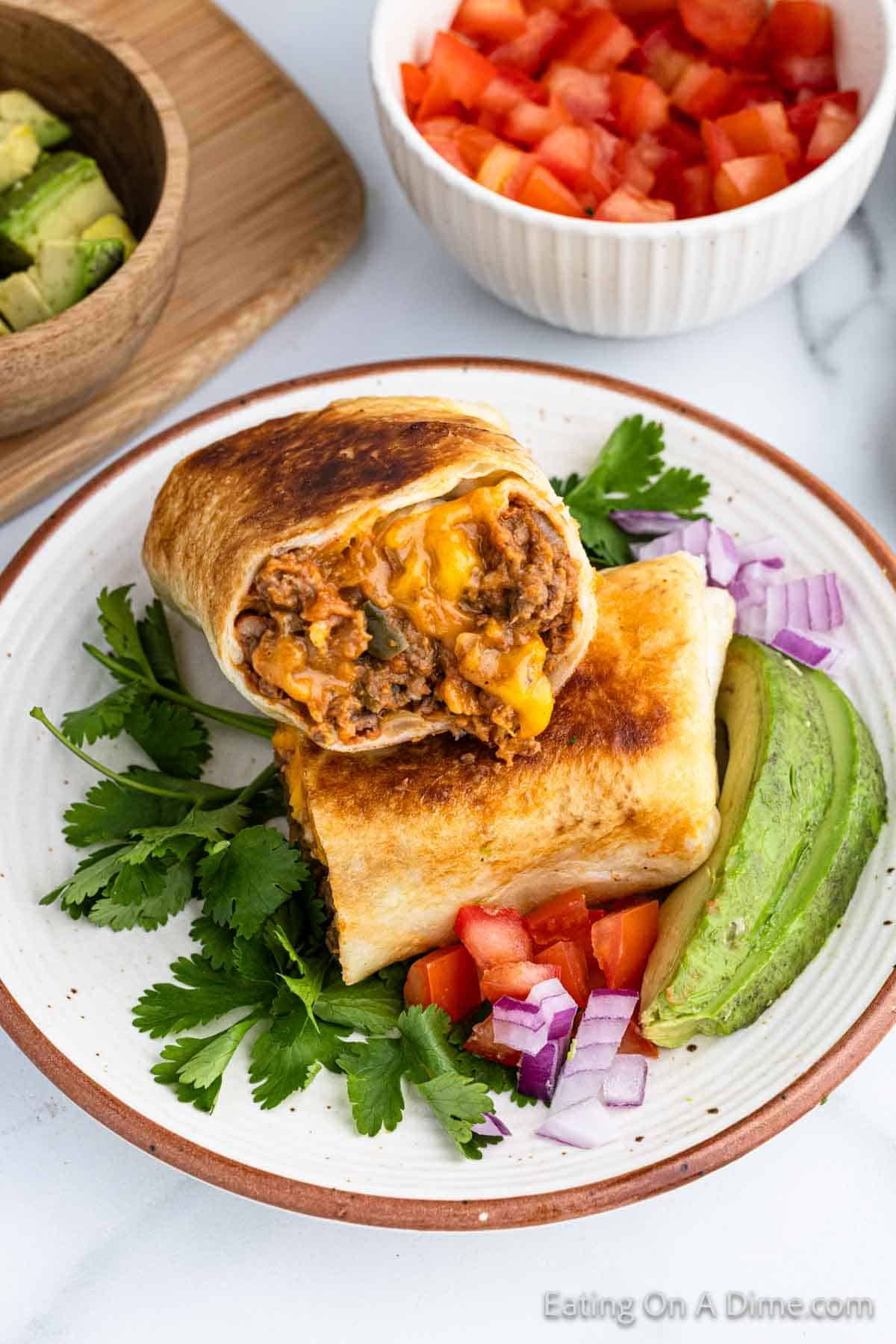 A beef chimichanga cut in half, revealing its filling of meat, cheese, and vegetables, is placed on a white plate with fresh cilantro, sliced avocado, diced tomatoes, and chopped red onions. Bowls with additional diced tomatoes and avocado pieces are in the background.