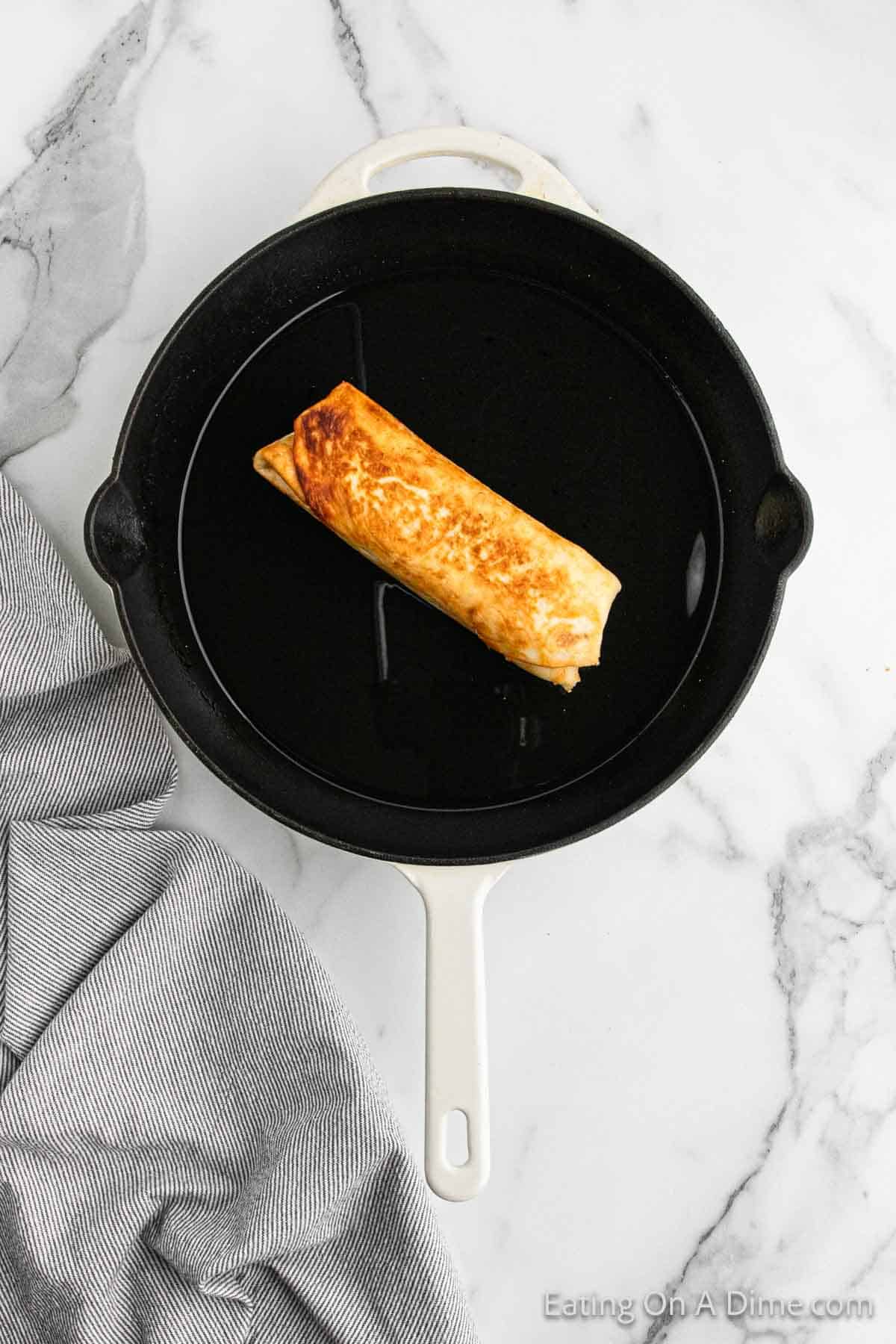 A single beef chimichanga is being cooked in a black cast iron skillet. The skillet is placed on a white marble countertop with a grey and white striped kitchen towel beside it. The chimichanga is partially browned, showcasing its crisp texture.