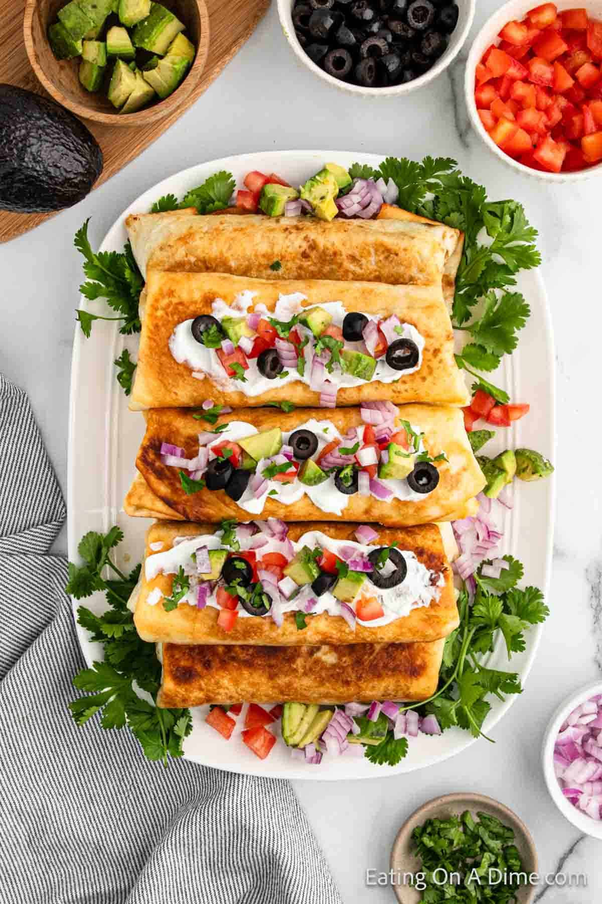 A white plate with three golden-brown beef chimichangas topped with sour cream, diced tomatoes, red onions, black olives, and chopped cilantro. The plate is surrounded by bowls of black olives, diced red peppers, and avocado chunks, with fresh parsley as garnish.