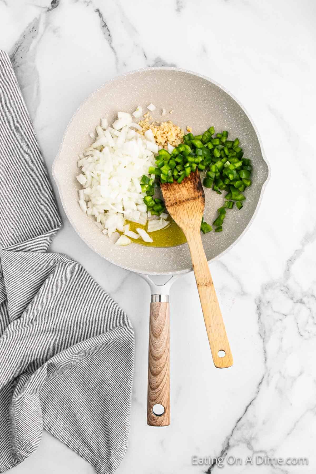A frying pan with chopped onions, minced garlic, and diced green bell peppers sits on a marble countertop, ready to make beef chimichangas. A wooden spatula rests in the pan, and a gray dish towel is draped nearby.