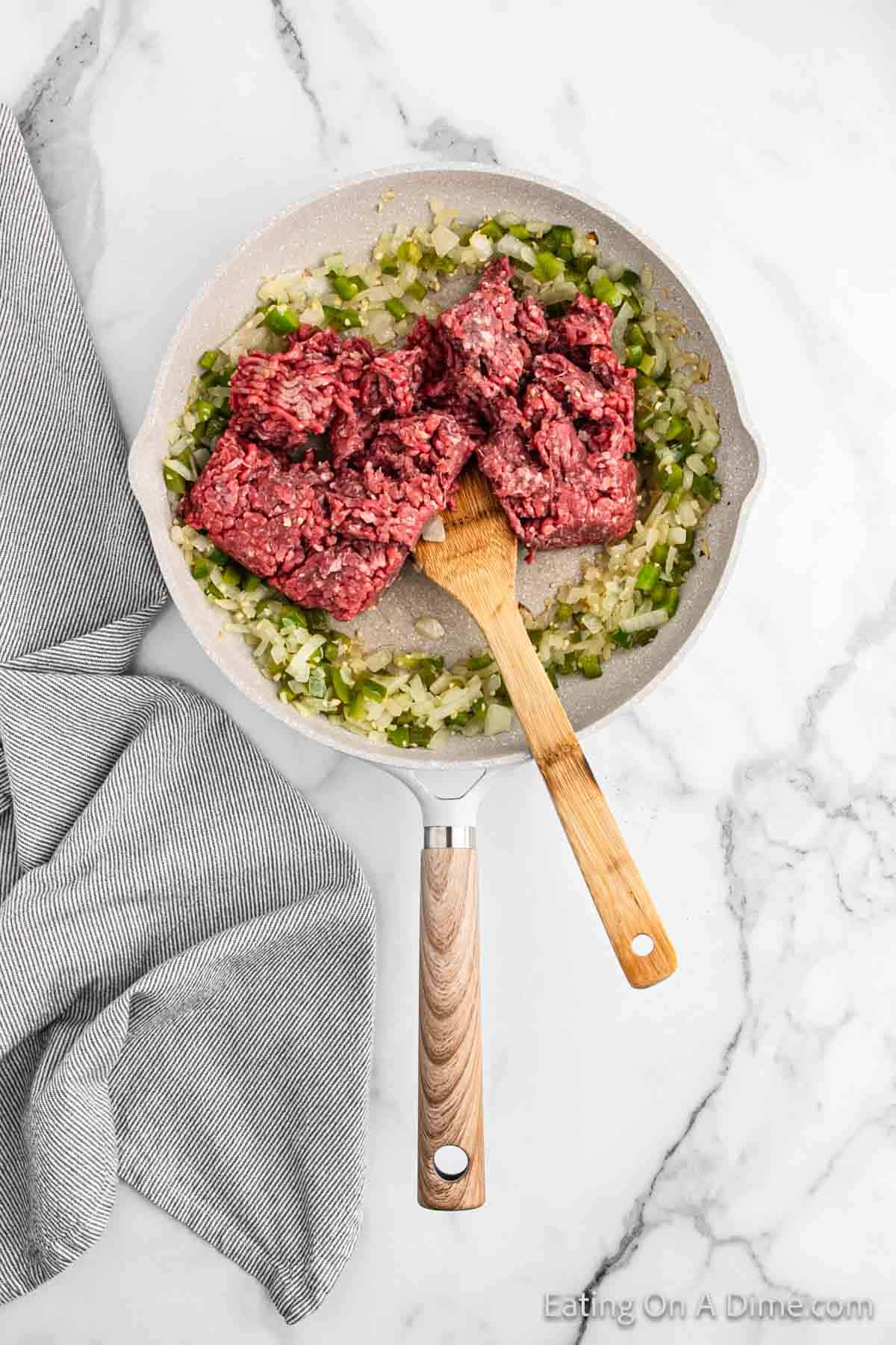 A frying pan with raw ground beef for Beef Chimichangas placed on top of sautéed onions and green peppers, being stirred with a wooden spatula. The pan is sitting on a marble countertop next to a folded gray-striped kitchen towel.