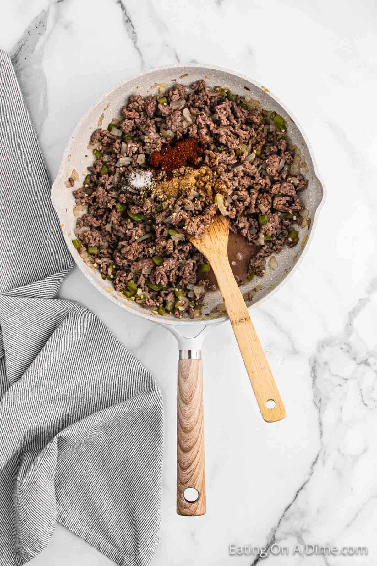 A skillet on a marble countertop contains cooked ground beef mixed with chopped onions and green bell peppers, reminiscent of beef chimichangas. Various spices are heaped on top of the mixture. A wooden spatula rests in the skillet, while a gray-and-white striped kitchen towel is placed beside it.