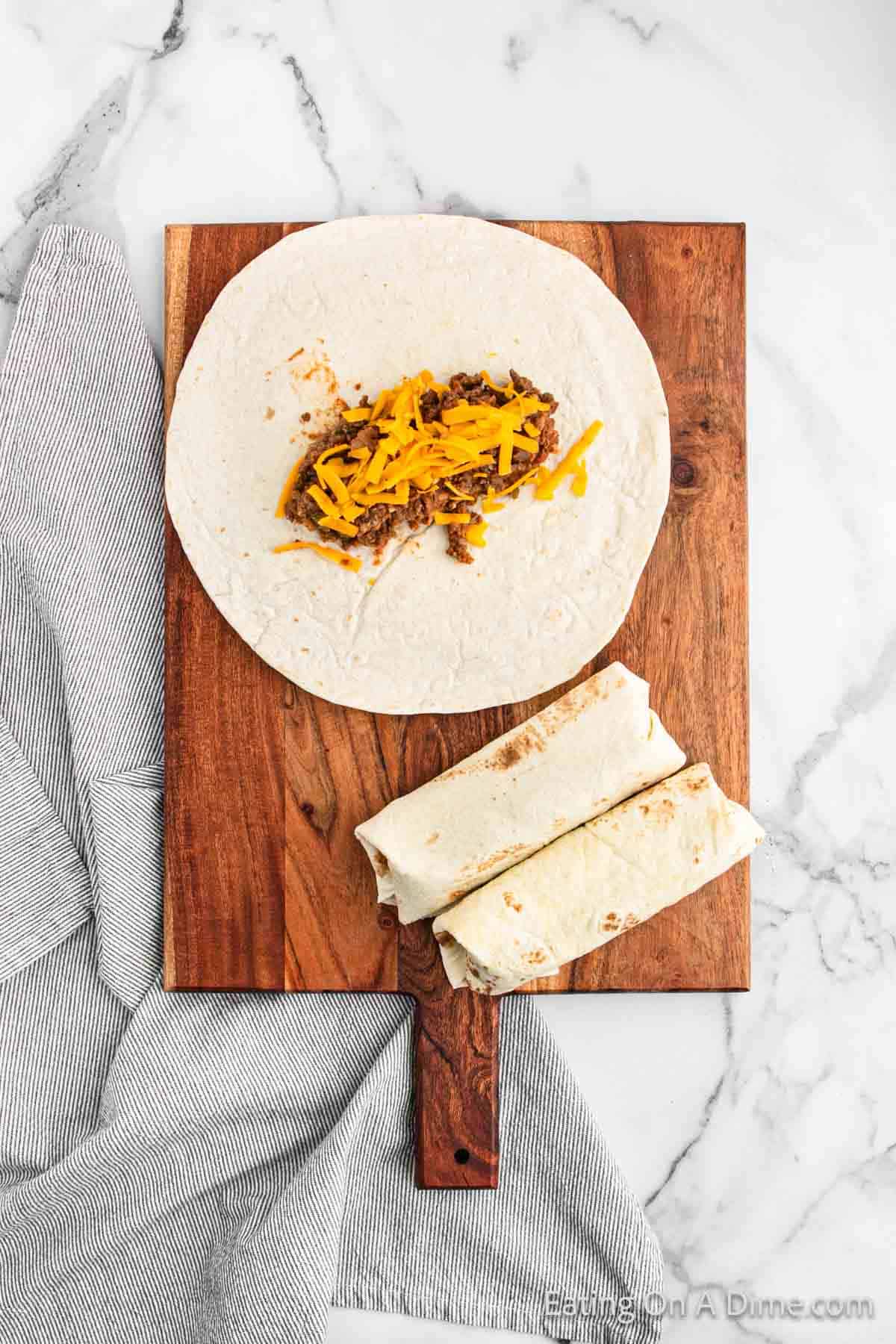 A wooden board rests on a marble surface, holding one open flour tortilla with ground beef and shredded cheddar cheese and two wrapped tortillas, poised to become delicious beef chimichangas. A gray-striped cloth is partially visible under the board.