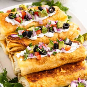 Golden brown beef chimichangas topped with sour cream, diced tomatoes, red onions, avocado, black olives, and cilantro, arranged on a white plate. Fresh cilantro leaves are scattered around the plate as garnish.