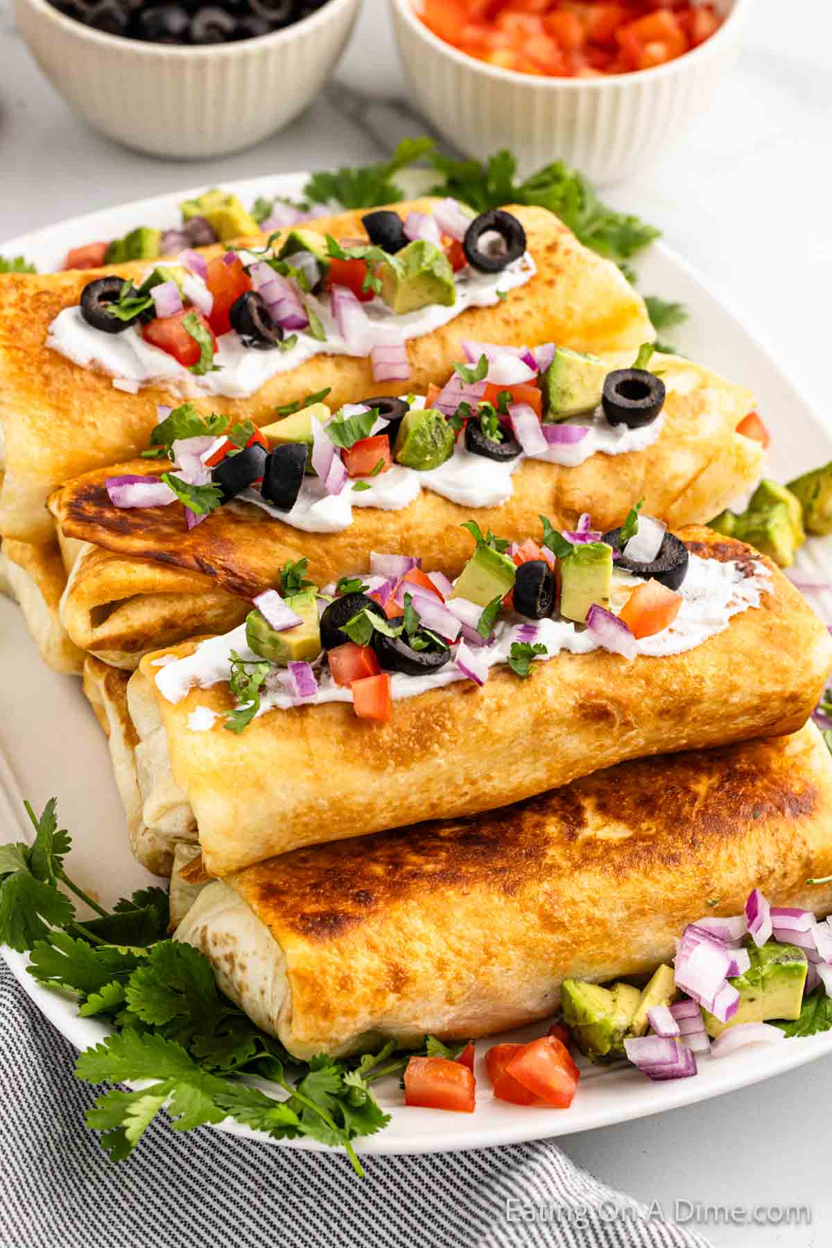 A plate of golden-brown beef chimichangas topped with sour cream, chopped tomatoes, red onions, black olives, and cilantro. Surrounding the plate are small bowls containing black olives and diced tomatoes. The vibrant ingredients add a splash of color to the dish.
