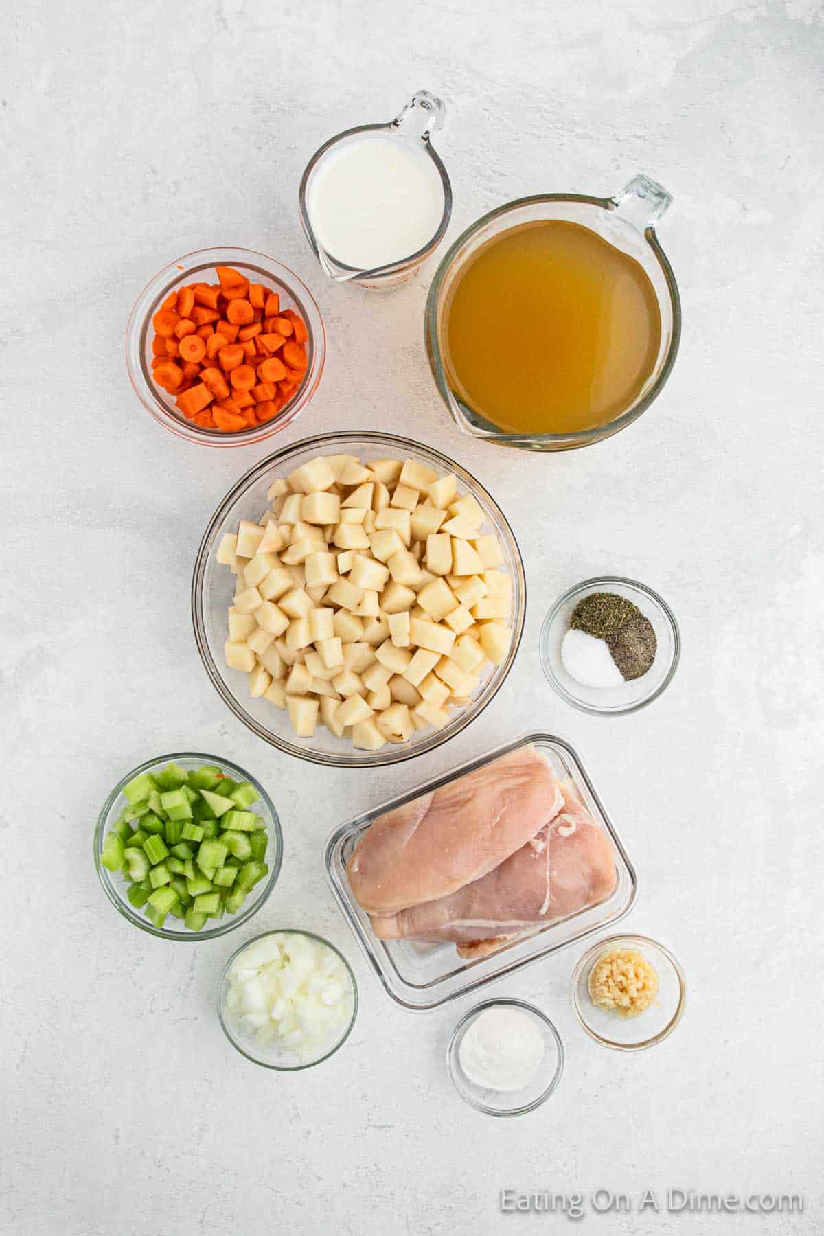 Ingredients for a crock pot potato soup with chicken are arranged on a white surface. They include diced potatoes, sliced carrots, chopped celery, chopped onion, raw chicken breast, minced garlic, salt, pepper, heavy cream, chicken broth, and butter.