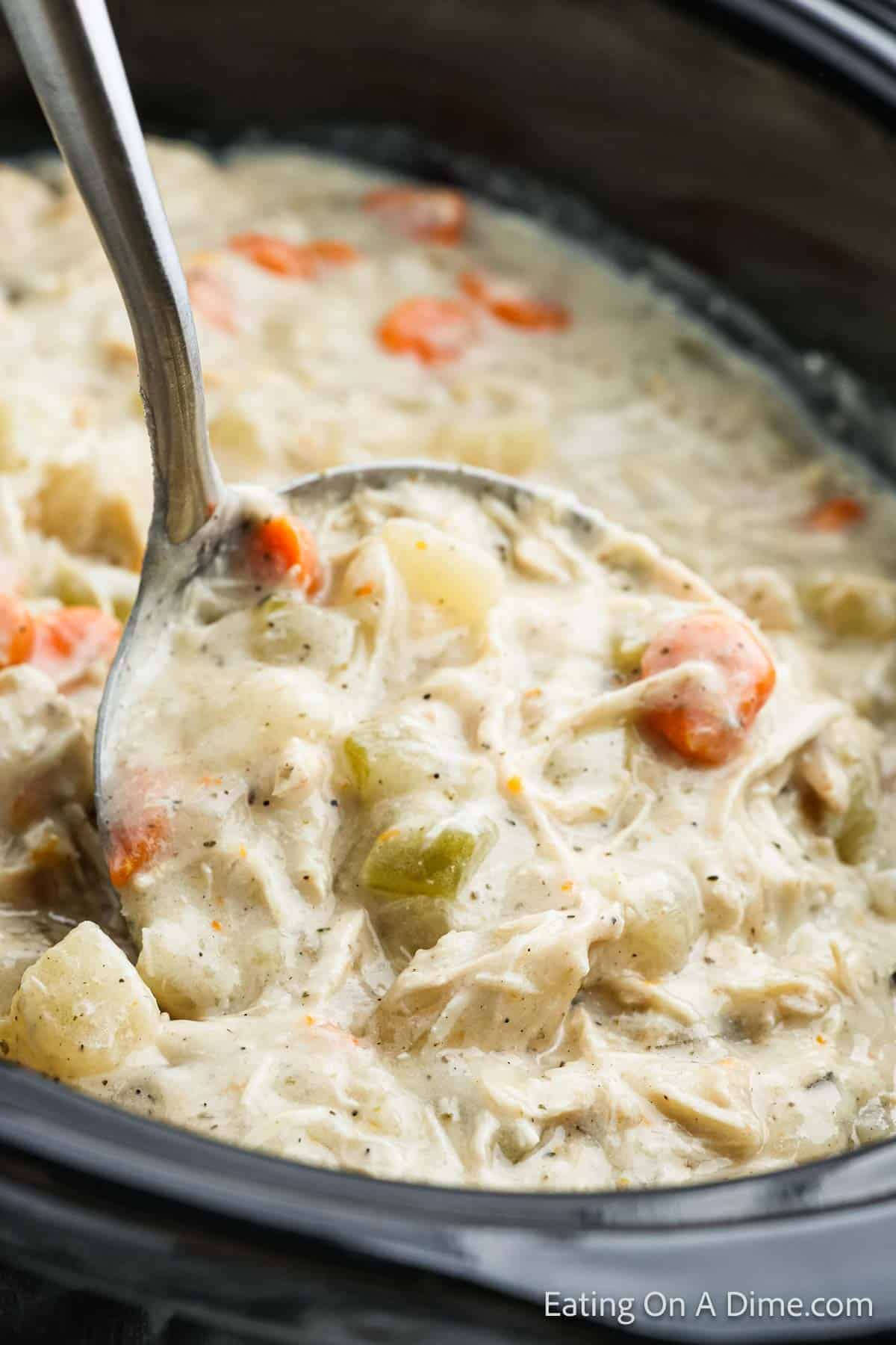 A ladle scoops creamy crock pot potato soup with chicken and vegetables from a slow cooker. The hearty mixture includes shredded chicken, chunks of potatoes, slices of carrots, and celery in a thick, savory broth. The image source is credited to Eating On A Dime at the bottom right.