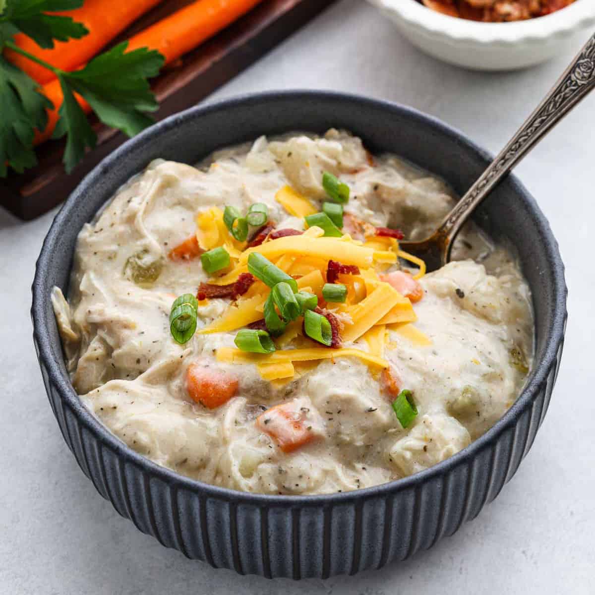 A bowl of creamy chicken pot pie soup, reminiscent of a savory crock pot potato soup with chicken, is garnished with shredded cheese, green onions, and bacon bits on a light-colored surface. A spoon rests inside the bowl while fresh carrots and parsley are visible in the background.