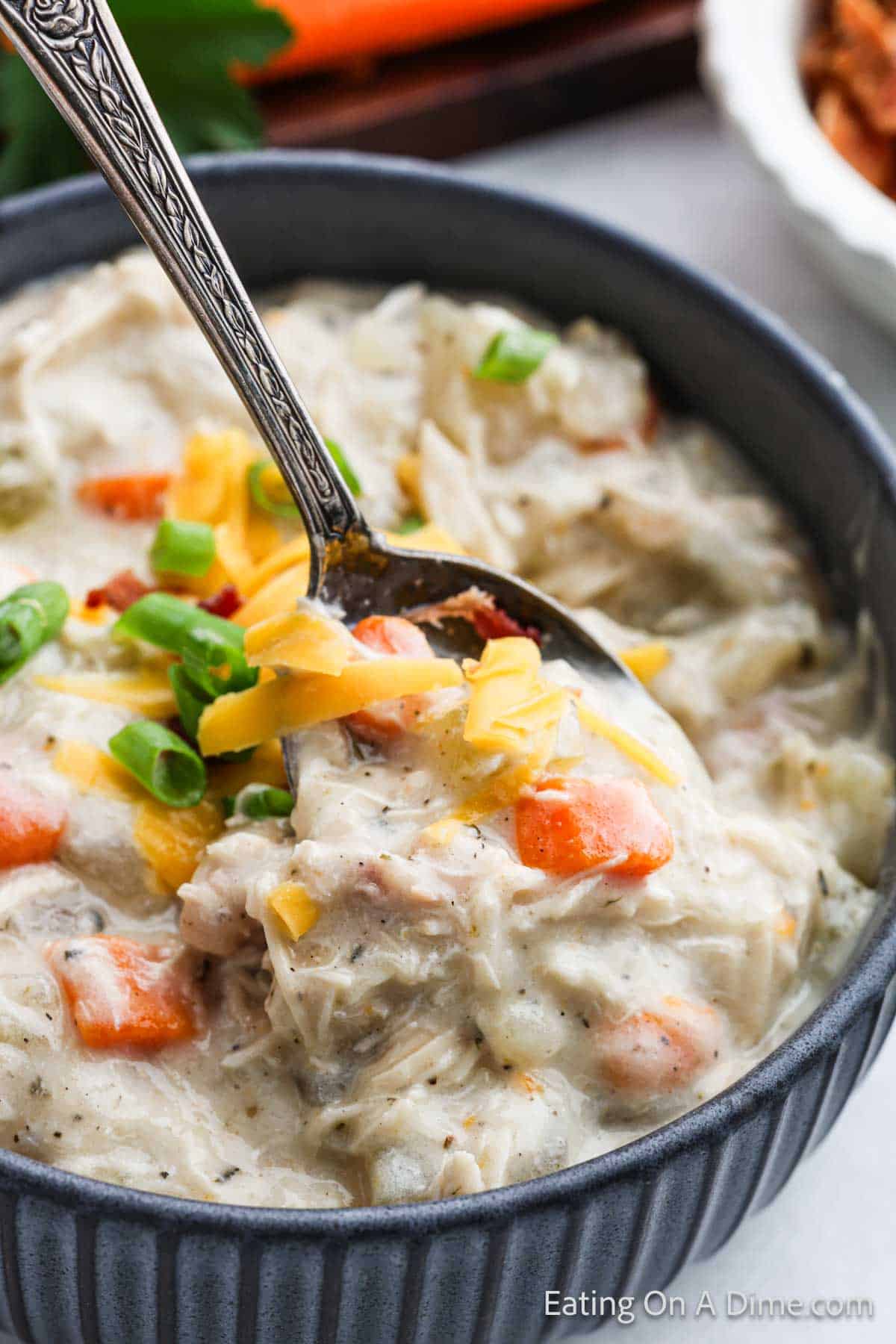 A close-up of a creamy chicken and rice soup served in a dark bowl, reminiscent of crock pot potato soup with chicken, garnished with chopped green onions and shredded cheese. The spoon, also in the bowl, holds a portion of the soup with chunks of chicken and carrots visible.