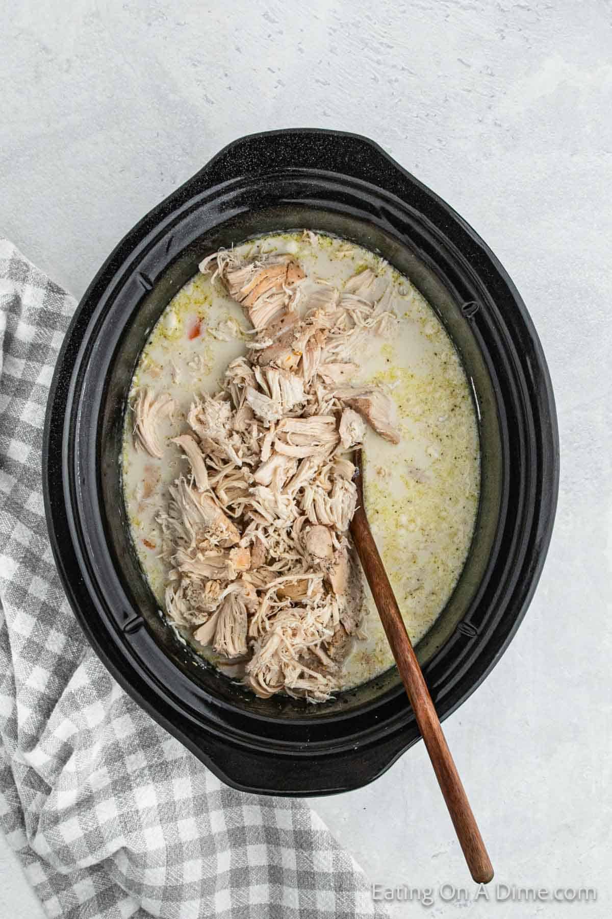 A slow cooker brimming with shredded chicken and creamy broth mixture, perfect for a crock pot potato soup with chicken. A wooden spoon rests inside the cooker, ready to serve. A grey and white checkered towel is placed beside the cooker on a light grey surface backdrop.