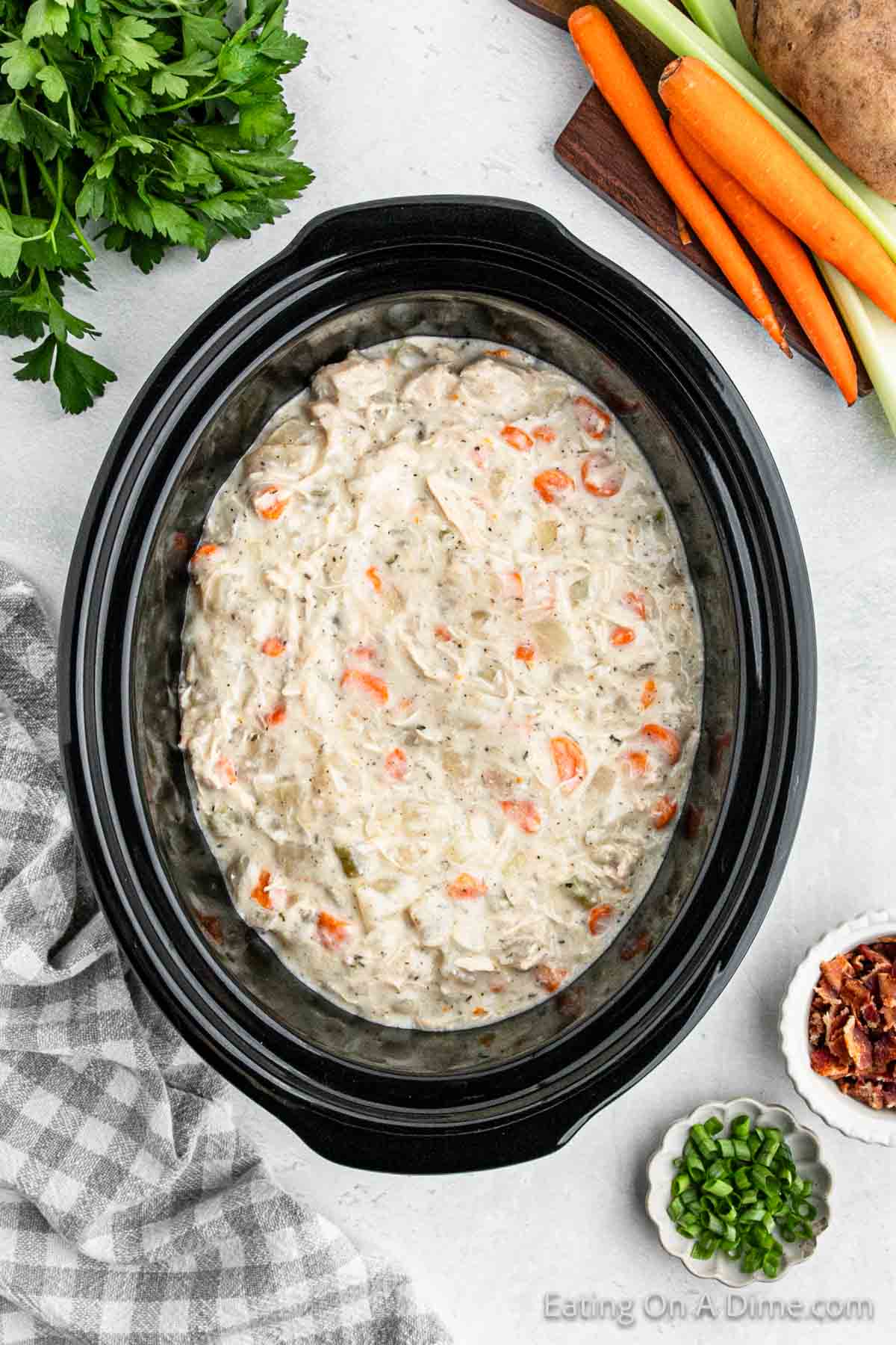 A black crock pot filled with creamy chicken and vegetables, featuring visible chunks of carrots and celery, sits on a light surface. Surrounding it are fresh parsley, a wooden board with carrots and celery, a potato, chopped green onions, and a small bowl of bacon bits—perfect for crock pot potato soup with chicken.