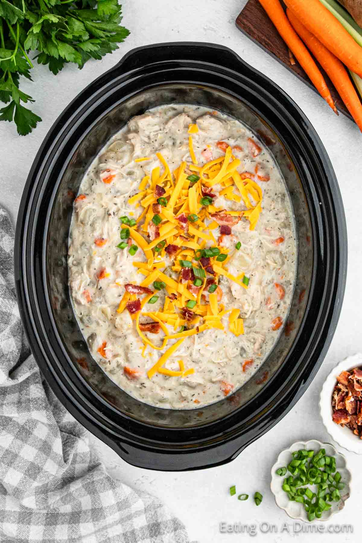 A crock pot bursting with creamy chicken and rice soup, garnished with shredded cheese, chopped green onions, and bacon bits. Surrounding the pot are fresh carrots, a bunch of parsley, a bowl of bacon bits, and a plate of sliced green onions.