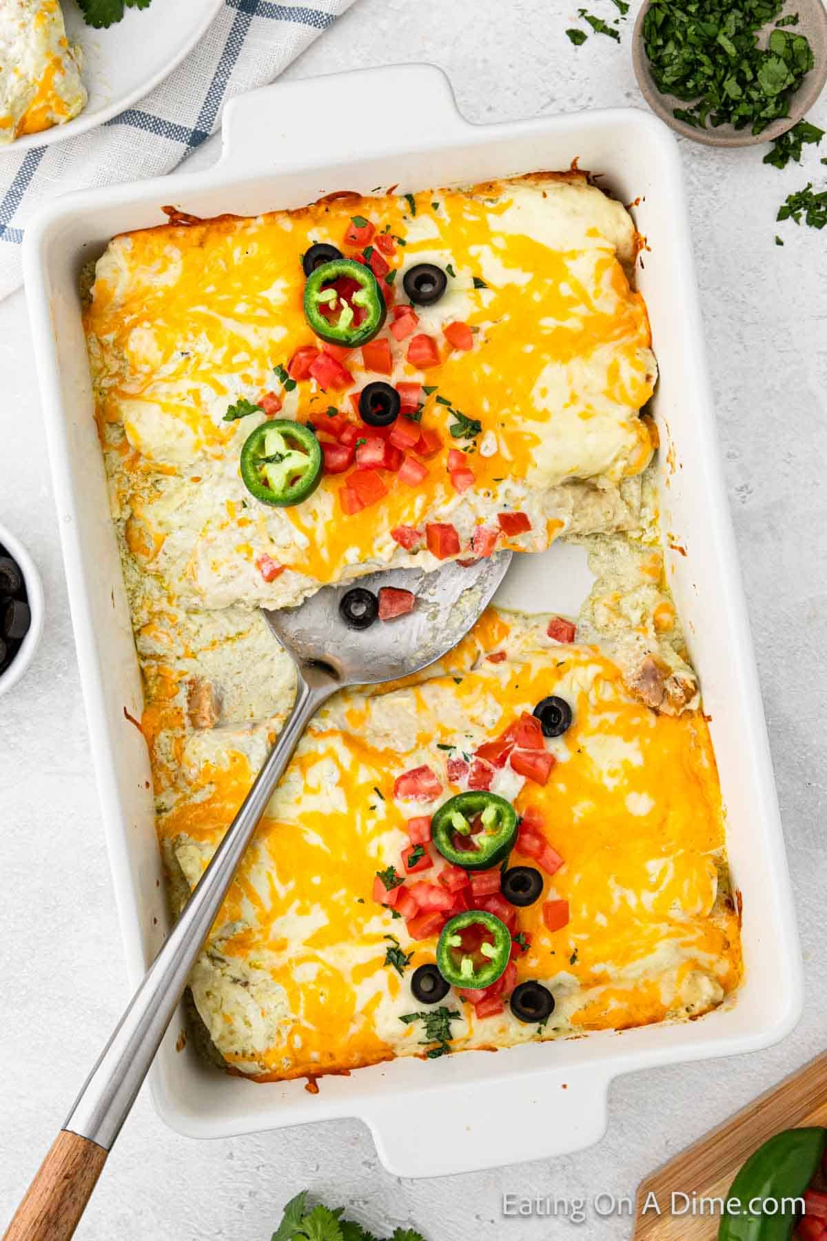 Chicken Enchiladas in a baking dish topped with diced tomatoes and slice jalapenos with a silver spatula ready to serve