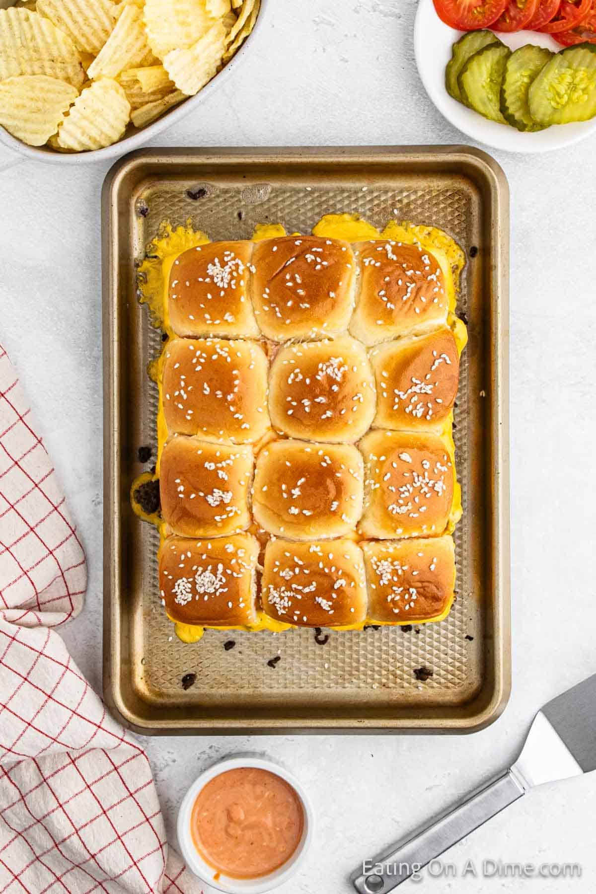 A tray of Hawaiian Roll Hamburger sliders topped with sesame seeds on a baking sheet, surrounded by a plate of potato chips, a dish with sliced tomatoes and pickles, a bowl of dipping sauce, and a checkered towel.