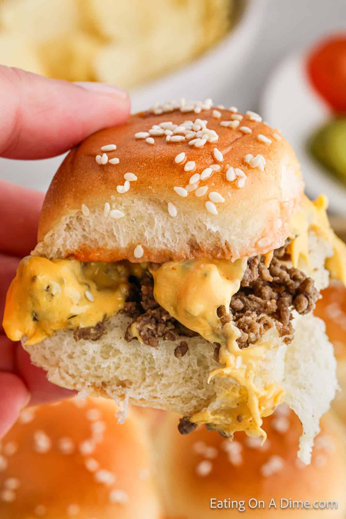 A hand holding a small cheeseburger slider with sesame seed bun. The slider, inspired by a Hawaiian Roll Hamburger recipe, is filled with ground beef and melted cheese. Blurred background shows another slider, vegetables, and a bowl of potato chips.