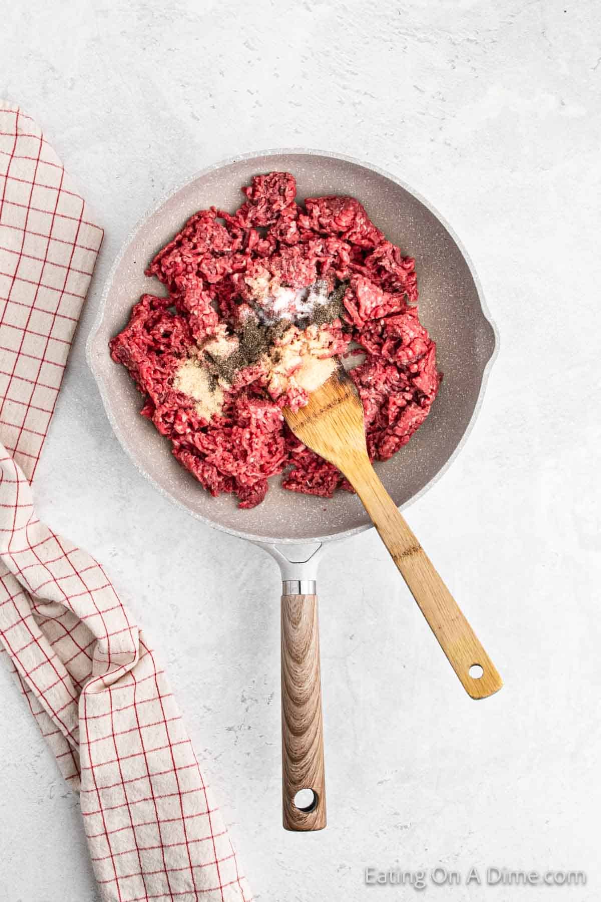 A skillet with raw ground beef and various seasonings, including minced garlic and ground black pepper, is being mixed with a wooden spatula. Perfect for a Hawaiian Roll Hamburger Recipe, a red and white checked kitchen towel lies beside the skillet on a light-colored surface.