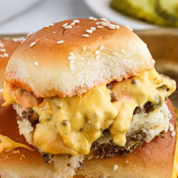 A close-up of a delicious cheeseburger slider. The slider has a fluffy Hawaiian Roll bun, a juicy beef patty, and is topped with melted cheese and sauce, dripping slightly over the sides. The background hints at a slice of pickle and another sandwich, perfect for your next hamburger recipe.