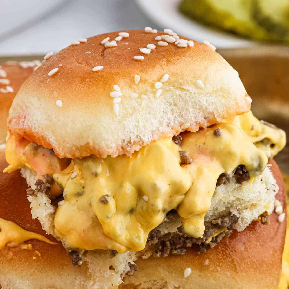 A close-up of a delicious cheeseburger slider. The slider has a fluffy Hawaiian Roll bun, a juicy beef patty, and is topped with melted cheese and sauce, dripping slightly over the sides. The background hints at a slice of pickle and another sandwich, perfect for your next hamburger recipe.
