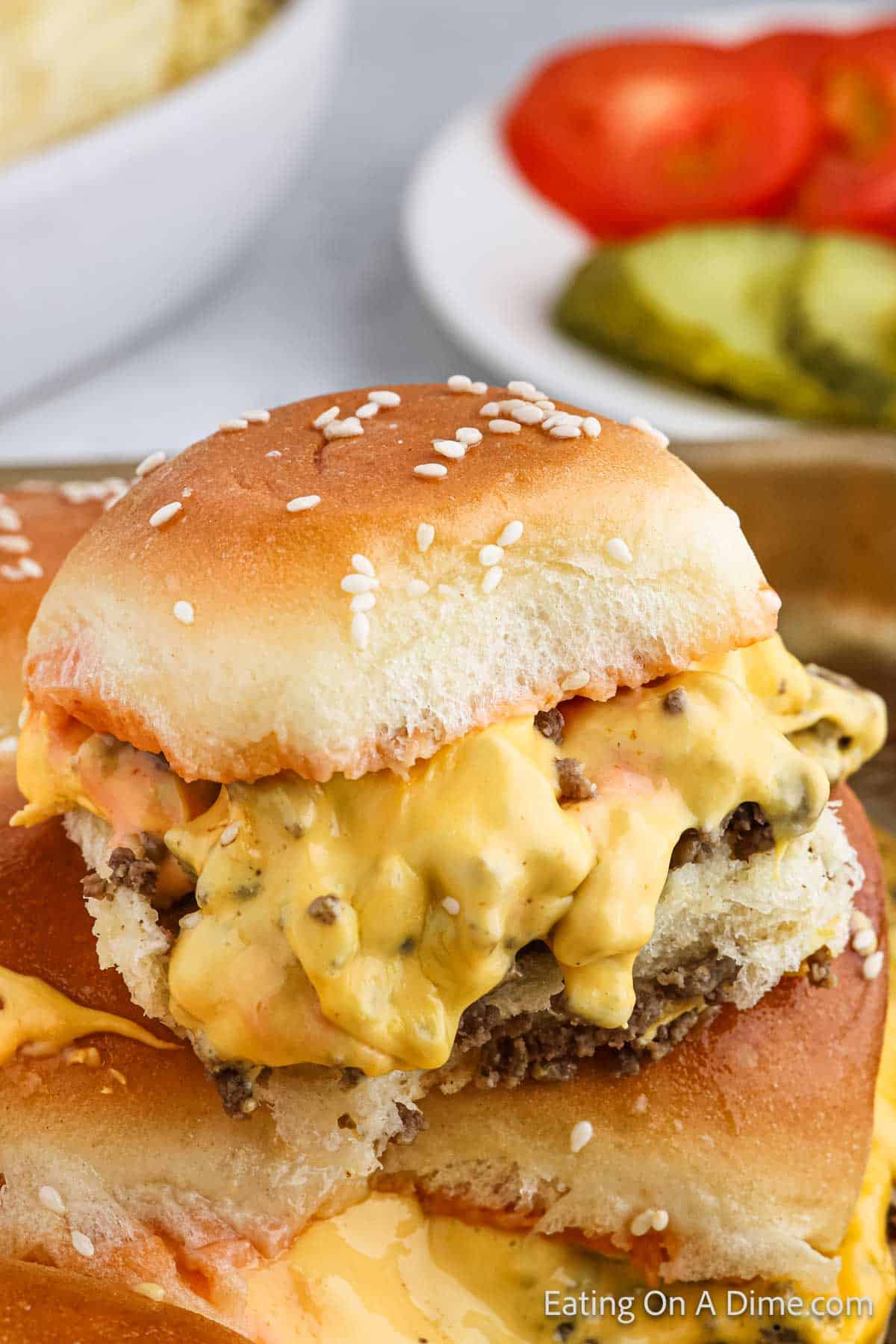 A close-up of a cheeseburger slider on a sesame seed bun. The slider is filled with melted cheese and a beef patty. In the background, there are sliced tomatoes and pickles on a plate, reminiscent of a delicious Hawaiian Roll Hamburger Recipe.