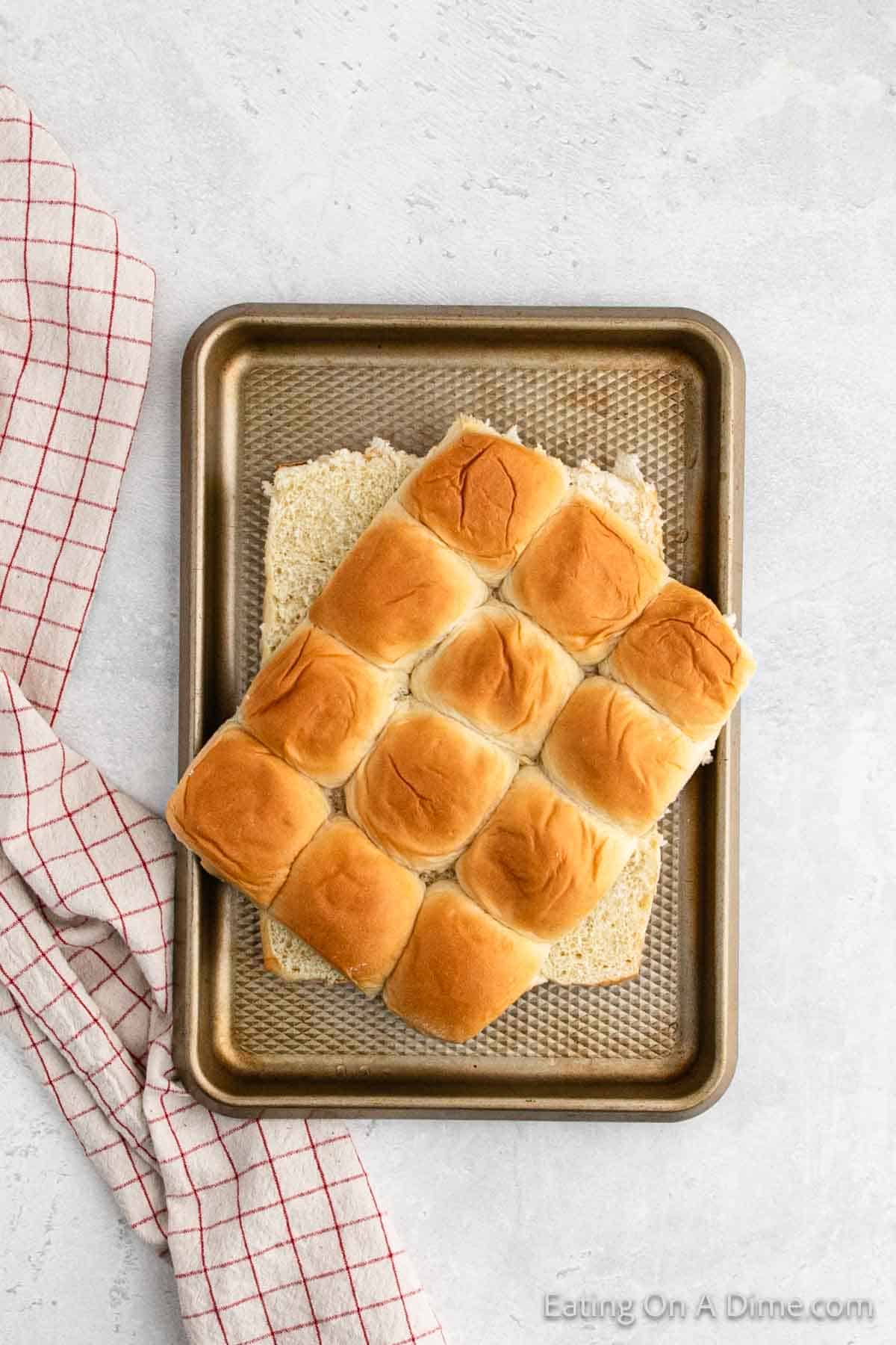 A baking sheet with a 12-roll sheet of golden brown Hawaiian rolls, perfect for your next Hawaiian Roll Hamburger Recipe, on a white countertop. A red and white checked cloth is placed beside the baking sheet.