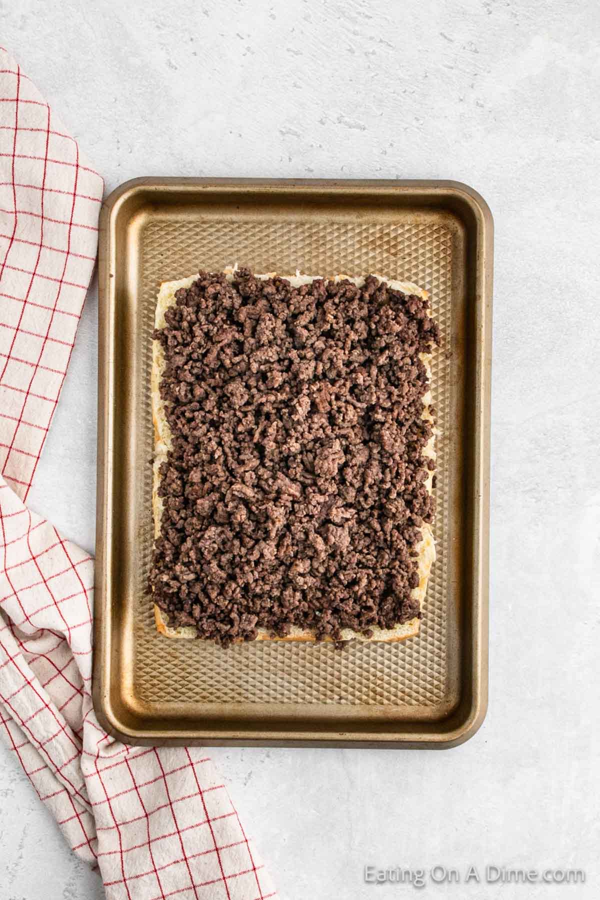 A metal baking tray with cooked, crumbled ground beef spread evenly on a piece of bread, reminiscent of a Hawaiian Roll Hamburger recipe. A white dish towel with red stripes is partially visible on the left side of the tray. The background is a light, textured surface.