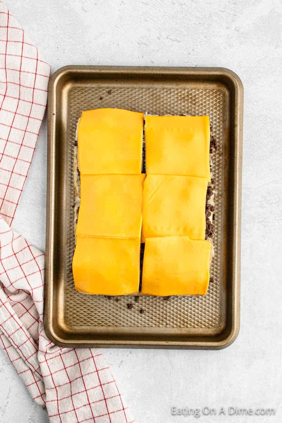 A baking tray with six square slices of cheddar cheese placed on top of cooked meat sits on a light gray surface, showcasing the deliciousness of a Hawaiian Roll Hamburger Recipe. A red and white checkered kitchen towel is partially visible on the left side of the tray. The corner of the tray reads "Eating On A Dime.com.